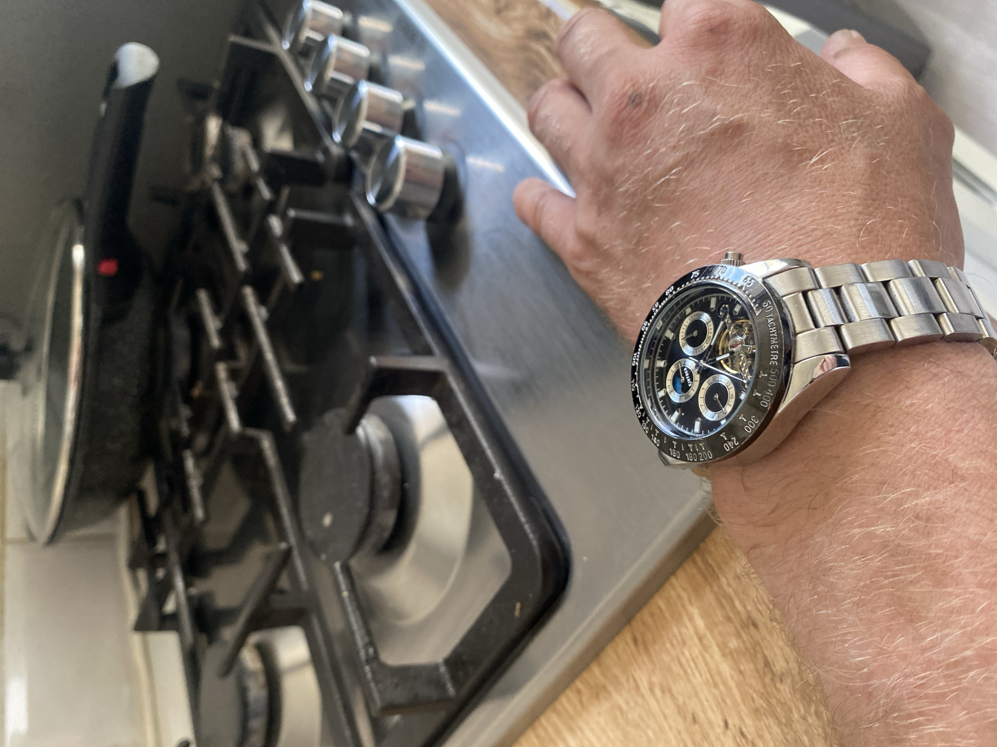 The image shows a close-up of a person's wrist wearing a silver-banded, black-faced wristwatch. The watch has multiple sub-dials and a tachymeter scale around the bezel, with visible text such as "TACHYMETRE S", "KINYUED", and numerical markings. The person's hand and arm, covered in light hair, are visible.
In the background, a stainless steel gas stovetop is prominently featured, with four burners and control knobs. To the left, part of a black cooking pot or pan is visible. The setting appears to be a kitchen, suggested by the stovetop and what looks like a wooden countertop. The lighting indicates it is daytime. The image does not convey any specific event or strong emotions, focusing instead on the watch and its immediate surroundings.