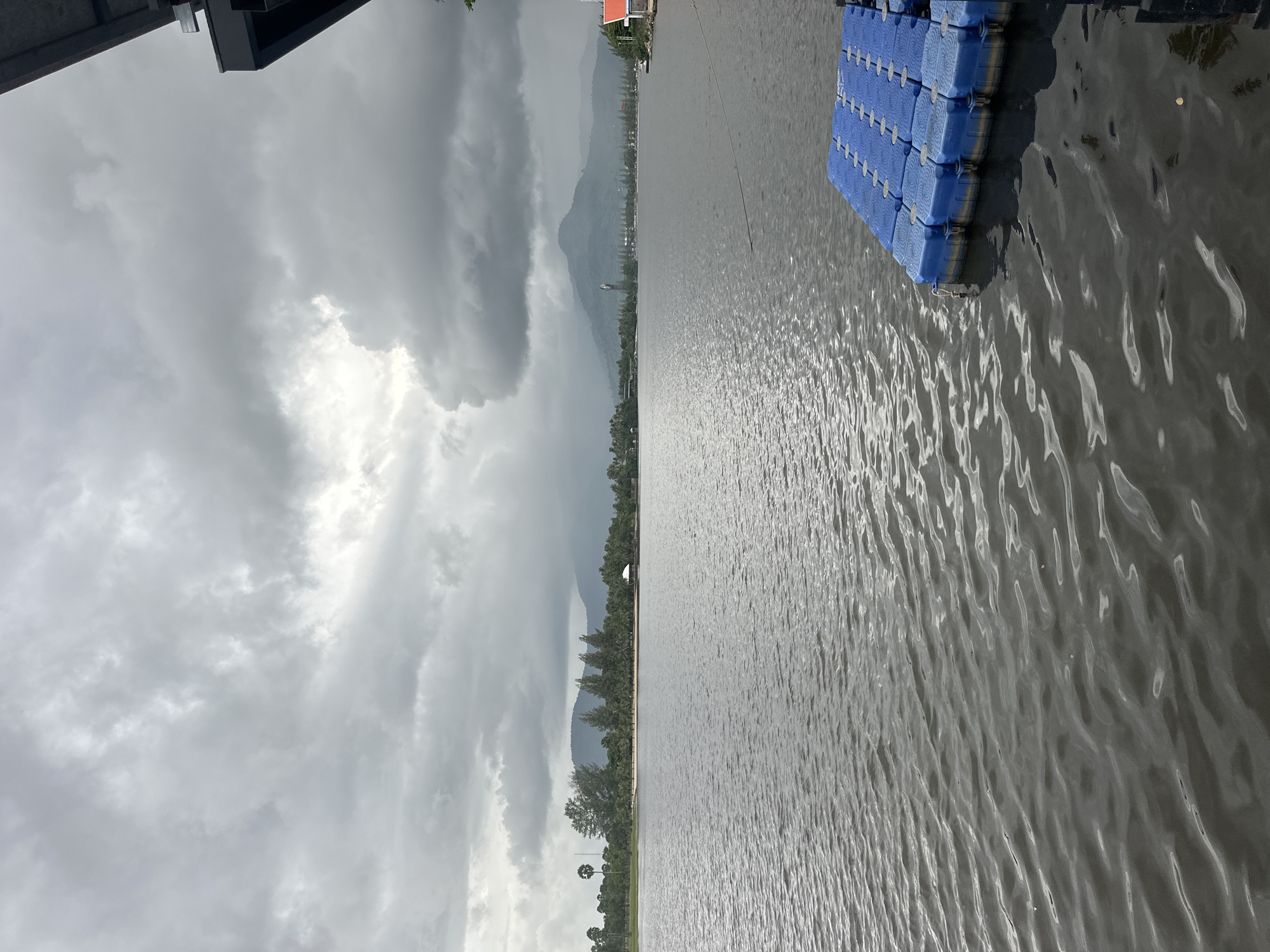 The image depicts a body of water with a cloudy sky overhead. In the foreground, a blue floating dock made of modular plastic cubes is visible on the right side. The water has a rippled surface. Across the water, a treeline with various trees, including palm trees, lines a sandy shore. In the distance, rolling hills and mountains are visible, with some structures, including what appears to be a building with a red roof, and possibly a tower or tall structure, along the shoreline and on the hills. The overall atmosphere is overcast and somewhat somber. This scene is consistent with locations along the riverbanks or coastal areas in or near Kampot, Cambodia, given the presence of lush vegetation, hills, and waterfront structures common to the region.