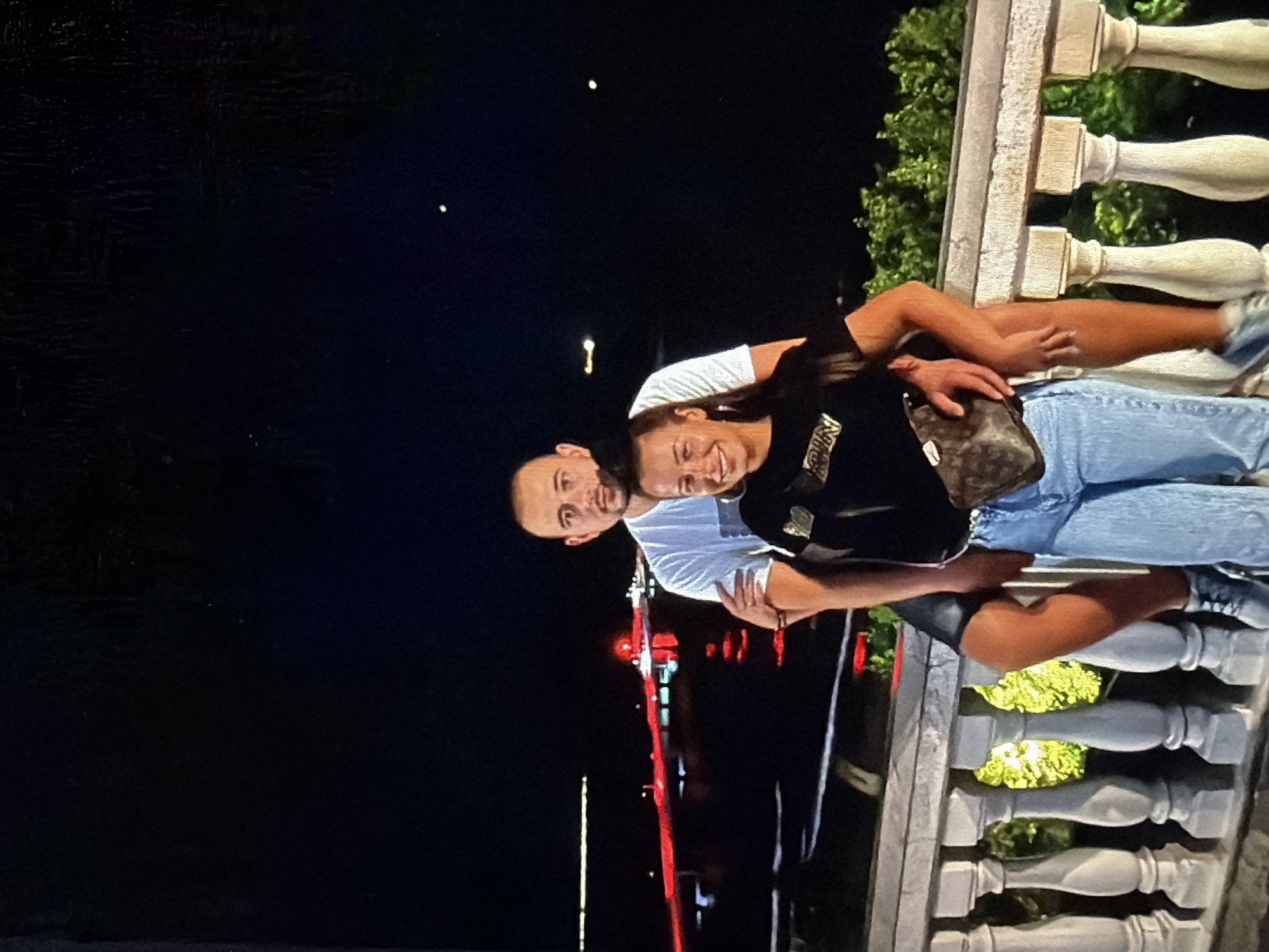A photograph captures a couple embracing on a balcony at night. The man, with a shaved head, wears a white T-shirt. The woman, with long dark hair, wears a black top and light blue jeans, with a patterned bag slung over her shoulder. They stand in front of a balustrade. In the background, streetlights and illuminated structures suggest an urban setting.
The photo was taken in Burgas, Bulgaria.