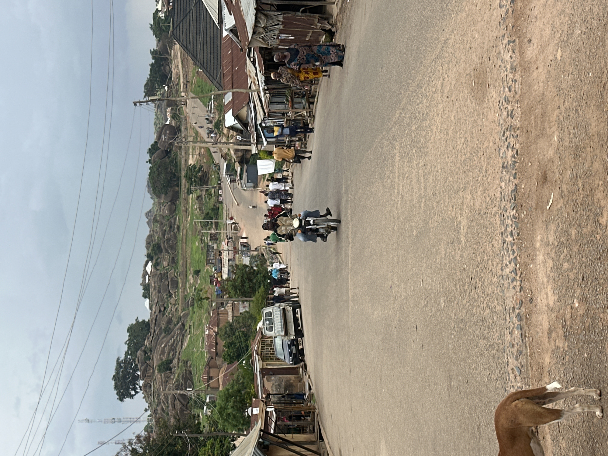 The image depicts a street scene in Jos, Nigeria. The street is paved and has people walking along the sides. A motorcycle is being driven on the street. Buildings with corrugated metal roofs line one side of the street. In the background, there is a rocky hill. There are power lines visible overhead. A dog is partially visible in the foreground. The sky is overcast.
