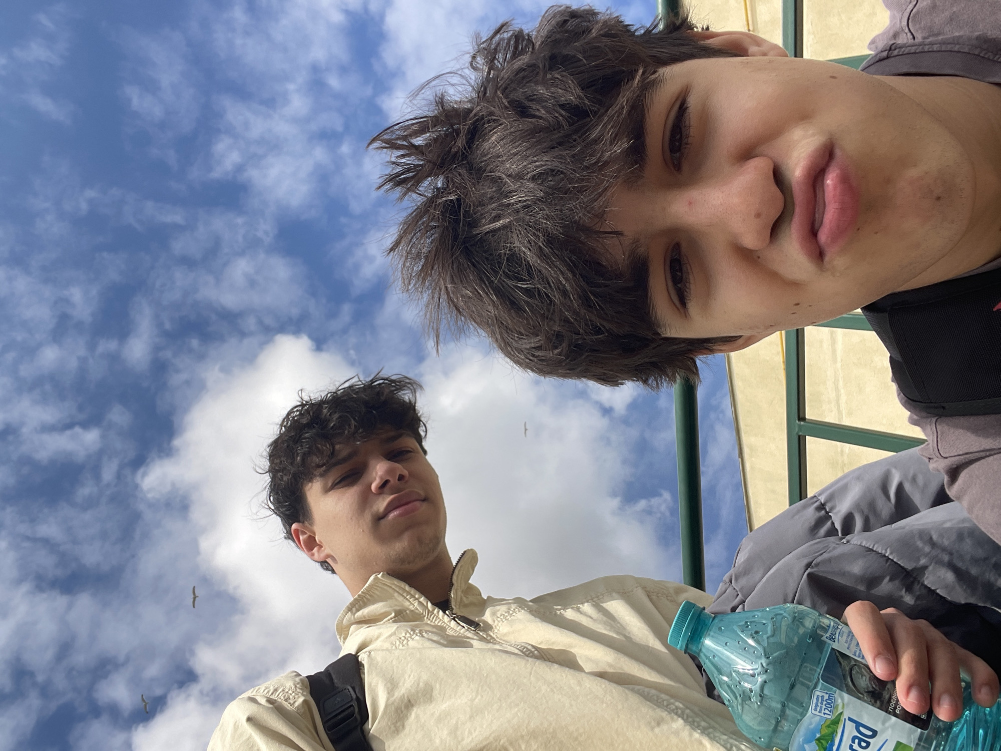Two young men are taking a selfie from a low angle against a bright, cloudy sky. The man in the foreground has dark, wavy hair and is wearing a light-colored jacket with a zipper. He also has a black backpack on his left shoulder and is holding a blue bottle of water. The second man, positioned slightly behind and above, has messy, dark hair. Some of the railing is green. The scene appears to be outdoors, and the presence of the water bottle and backpack suggests they might be tourists or travelers. Based on the architecture visible, it is possible the location is Varna, Bulgaria. Two birds can also be seen in the sky.