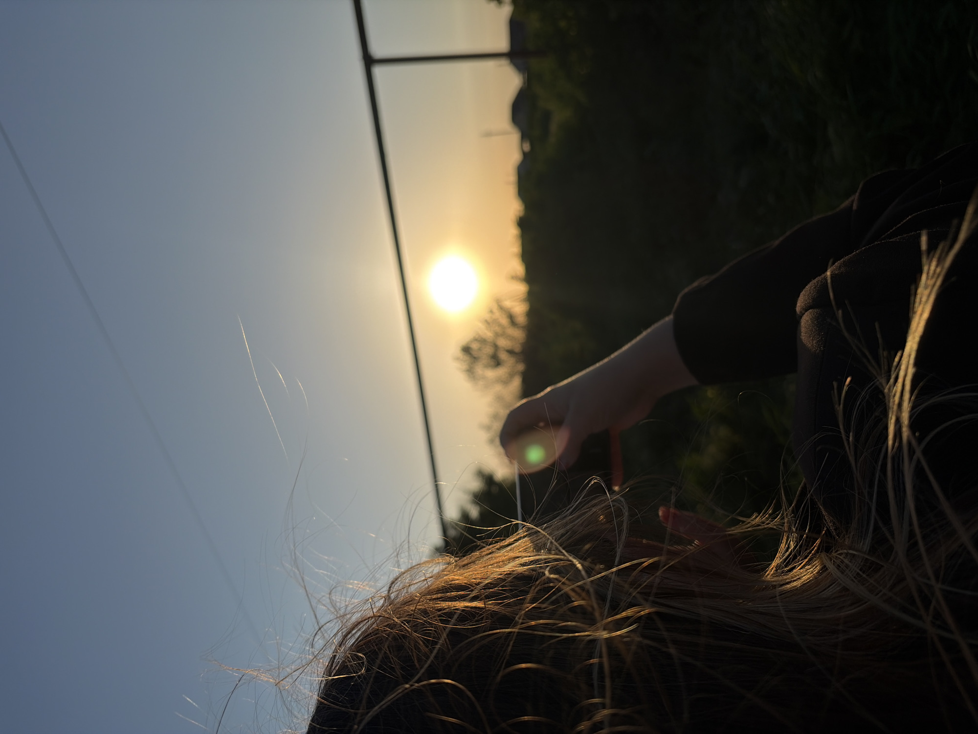 The image shows a low-angle view of a person with blonde hair and a black top, presumably in Novopokrovskaya, Russia. The person's hand is raised near their head, seemingly touching a strand of hair that reflects the sunlight. A bright sun is visible in the background, along with dark green foliage and a fence structure. The sky transitions from blue to a light, sunset-like color. The photograph seems to be taken during golden hour.