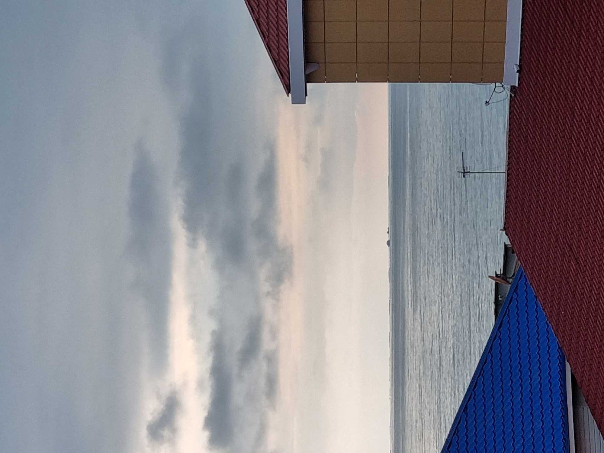 This image depicts a view of the sea from what appears to be a building or structure in Sochi, Russia. The image includes sections of rooftops with red, yellow, and blue roofing tiles. The sea is visible in the center of the image with a grey sky behind it, indicating either early morning or evening hours. There's an antenna visible above the blue rooftop on the right side of the image.