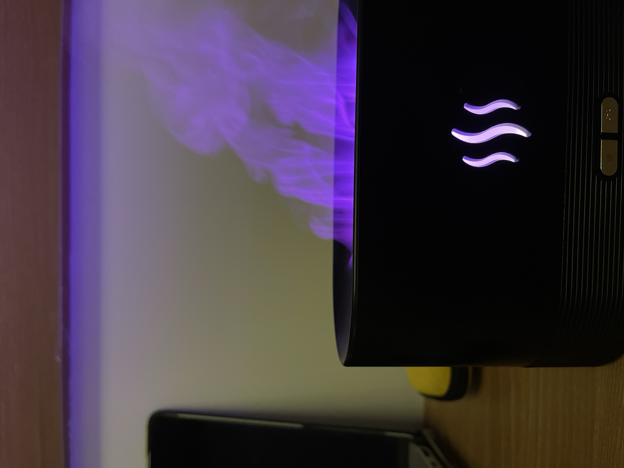 The image shows a close-up of a black aroma diffuser with purple mist emanating from its top. The diffuser has a stylized flame or steam design etched into its front, illuminated by purple light. Below the design are two small buttons. The diffuser is placed on a wooden surface, and to the left, a portion of a laptop and a yellow object are visible. In the background, a white wall is illuminated by a purple light source from above. The scene is indoors and appears to be a domestic or office setting. There is no indication of the city of Da Nang, Vietnam, or any specific location within it.