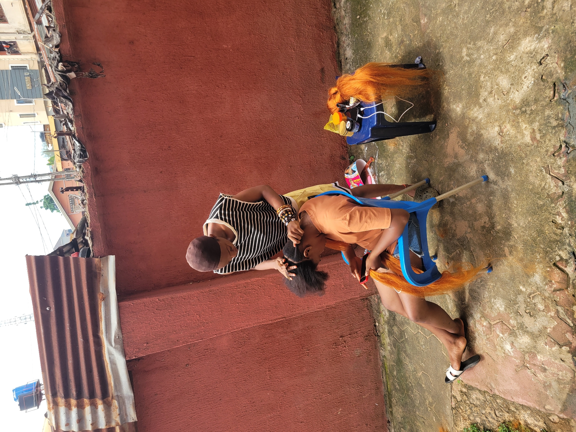Two individuals are engaged in braiding hair outdoors. The person on the left, wearing a black and white striped shirt and a brown cap, is actively braiding the hair of the person on the right. The person on the right, dressed in an orange t-shirt, is seated on a blue chair and has long, bright orange hair extensions being incorporated into their natural hair.
The scene is set against a reddish-brown textured wall. The ground appears to be a mix of concrete and possibly paving stones, with some areas showing moss or discoloration. To the left of the frame, a corrugated metal roof structure is visible, and further in the background, there are glimpses of buildings and possibly a water tank.
On a blue stand to the right of the seated person, a bundle of orange hair extensions is placed, along with what appears to be a small basket or container holding other hair accessories. A white electrical cord is also visible connected to the stand. The overall lighting suggests it is daytime, possibly late morning or early afternoon, with bright but diffused light, indicating a possibly overcast or generally bright day. The activity appears to be a casual, everyday event, with no strong emotional cues evident. There is no visible text in the image.