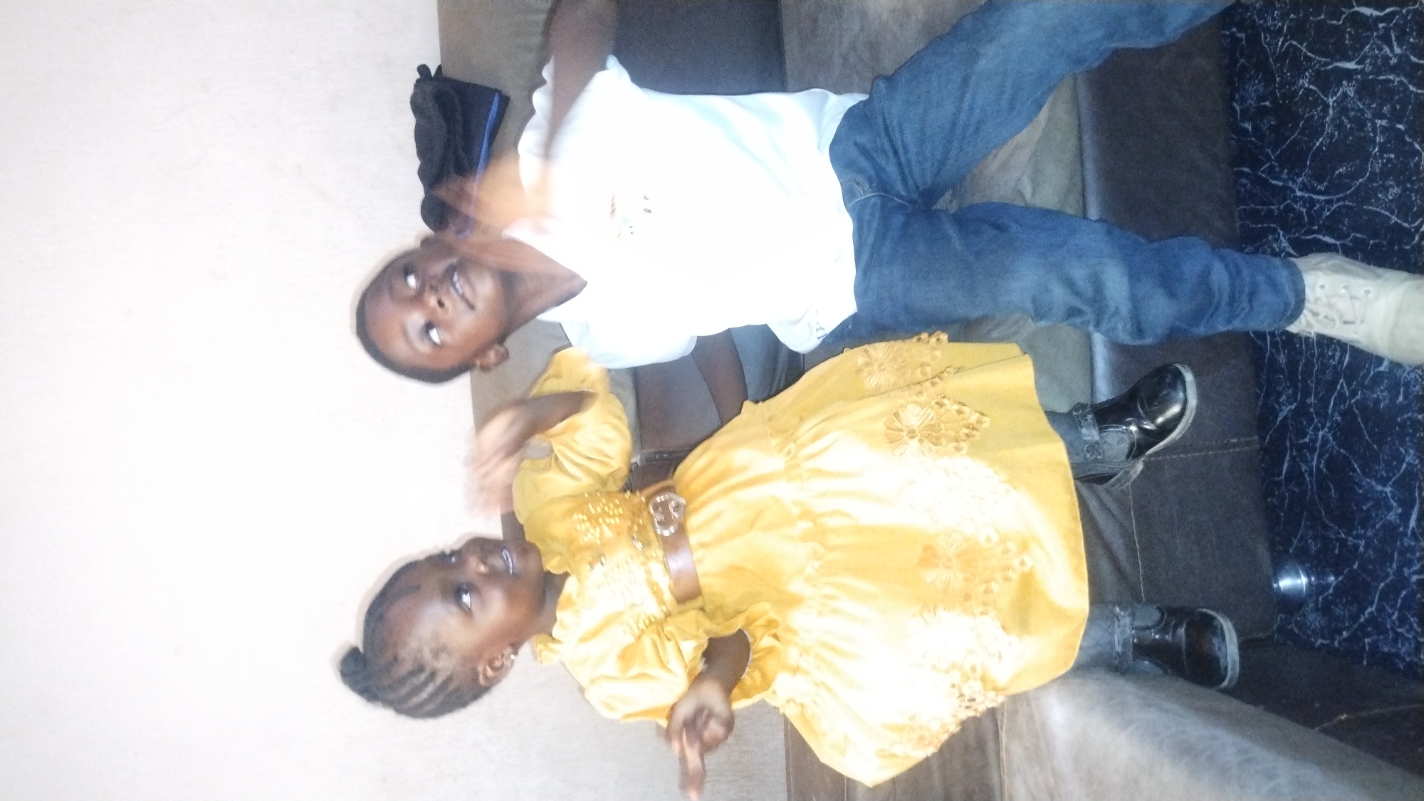 Two children are photographed indoors. A boy is wearing a white t-shirt and blue jeans, with off-white shoes on one foot and black shoes on the other. A girl is wearing a yellow dress with a brown belt and black shoes. The environment appears to be a casual living space with dark-colored furniture. There is a black bag to the left of the boy. The image was likely taken in Jos, Nigeria. The children are smiling.