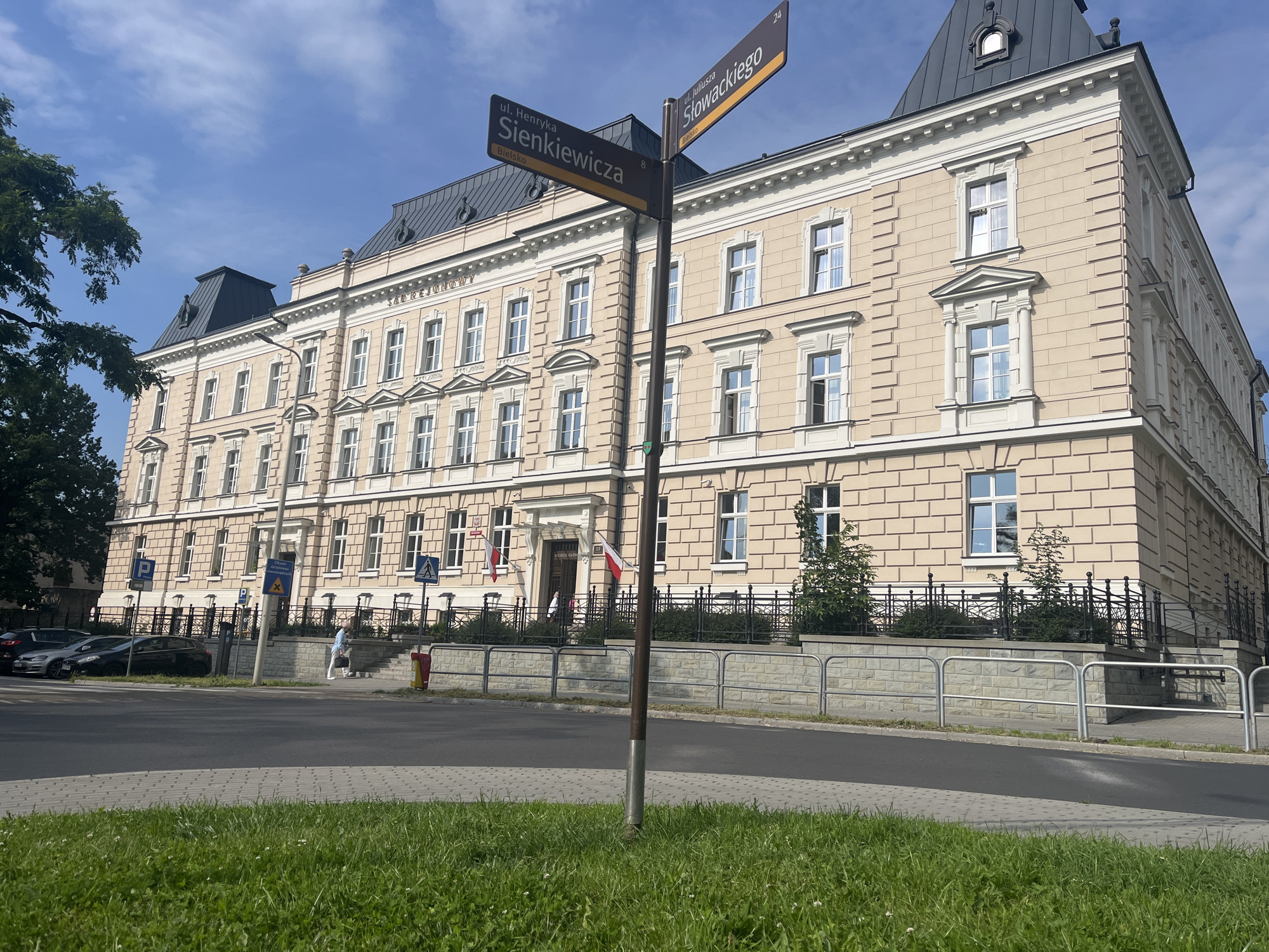 A street sign indicates the intersection of ul. Henryka Sienkiewicza and ul. Juliusza Słowackiego in Bielsko. The sign features numbers 8 and 24. The scene is dominated by a large, light-tan building with a dark, sloped roof. The building has a classical architectural style with numerous windows, decorative trim, and a grand entrance. Two Polish flags are displayed near the entrance. In front of the building, there is a black metal fence and a stone retaining wall. Several cars are parked along the street. A pedestrian, dressed in light blue and white, walks on the sidewalk. The foreground shows a grassy area and a paved walkway. The sky is blue with scattered white clouds.