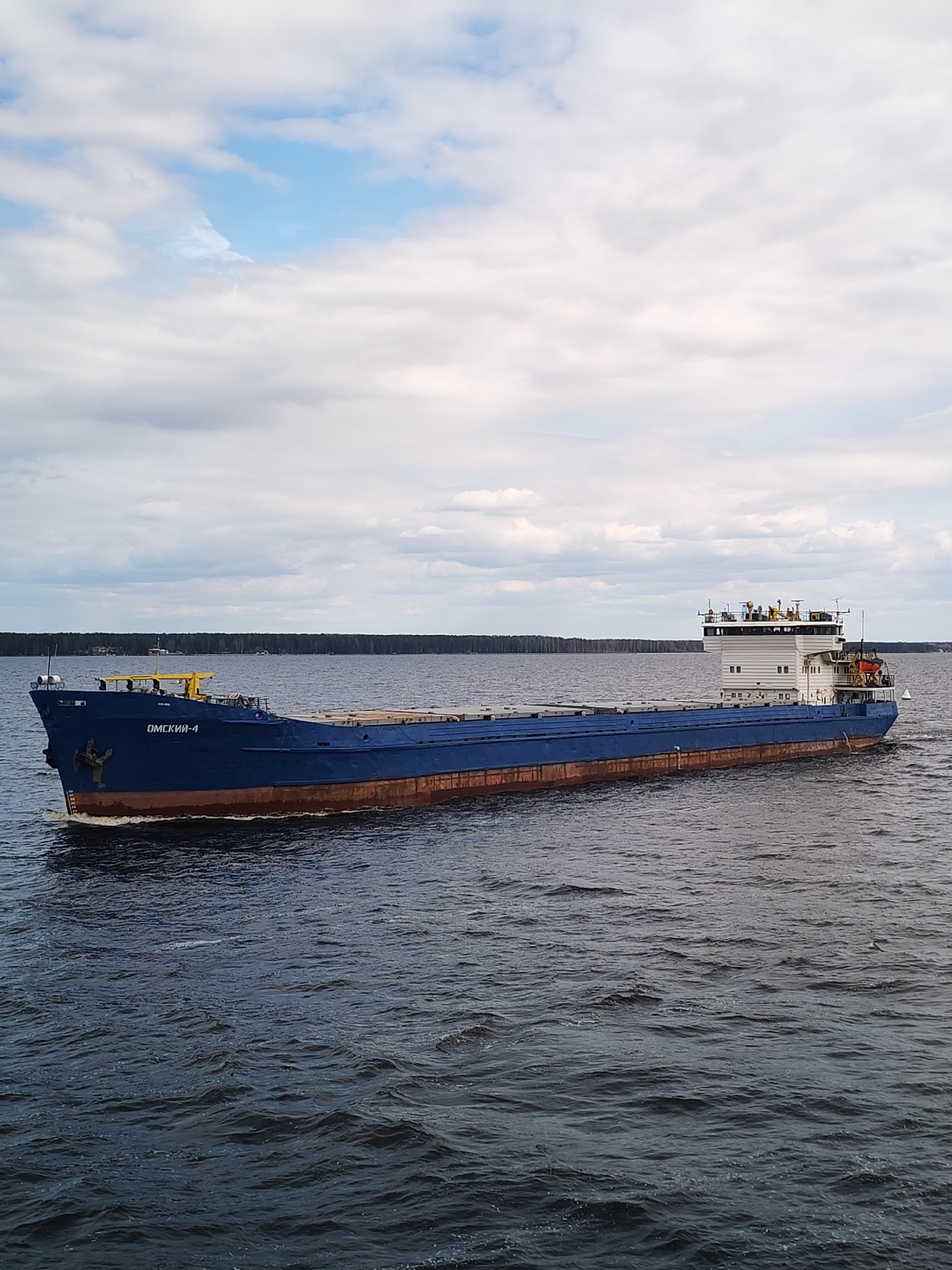 A blue cargo ship labeled "OMCKHH-4" is moving through a body of water. The ship has a brown hull and a white superstructure. The water is dark and rippled. In the background, there is a treeline and a cloudy sky. The location is difficult to identify precisely without additional information.