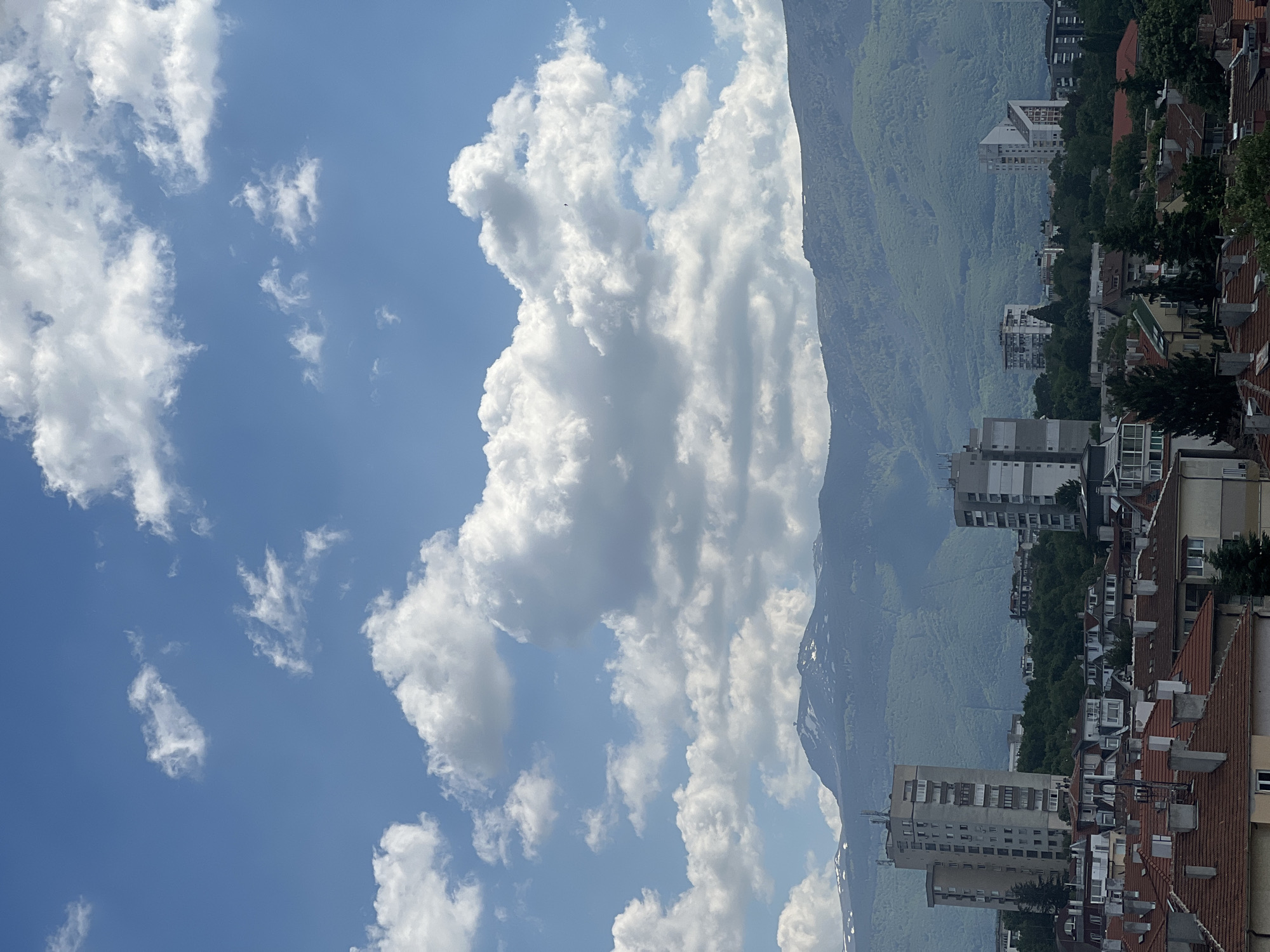 The image shows a view of Sofia, Bulgaria, taken at an upward angle, incorporating the sky, a mountain, and the city's buildings. The sky occupies the left portion of the frame, filled with scattered white cumulus clouds against a blue background. To the right, a mountain range extends vertically, covered in lush green vegetation. The foothills gradually transition into the urban landscape, with buildings nestled among trees. The architecture features a mix of residential buildings and taller apartment complexes, with visible rooftops and facades. The scene captures a serene, daytime atmosphere under clear weather conditions.