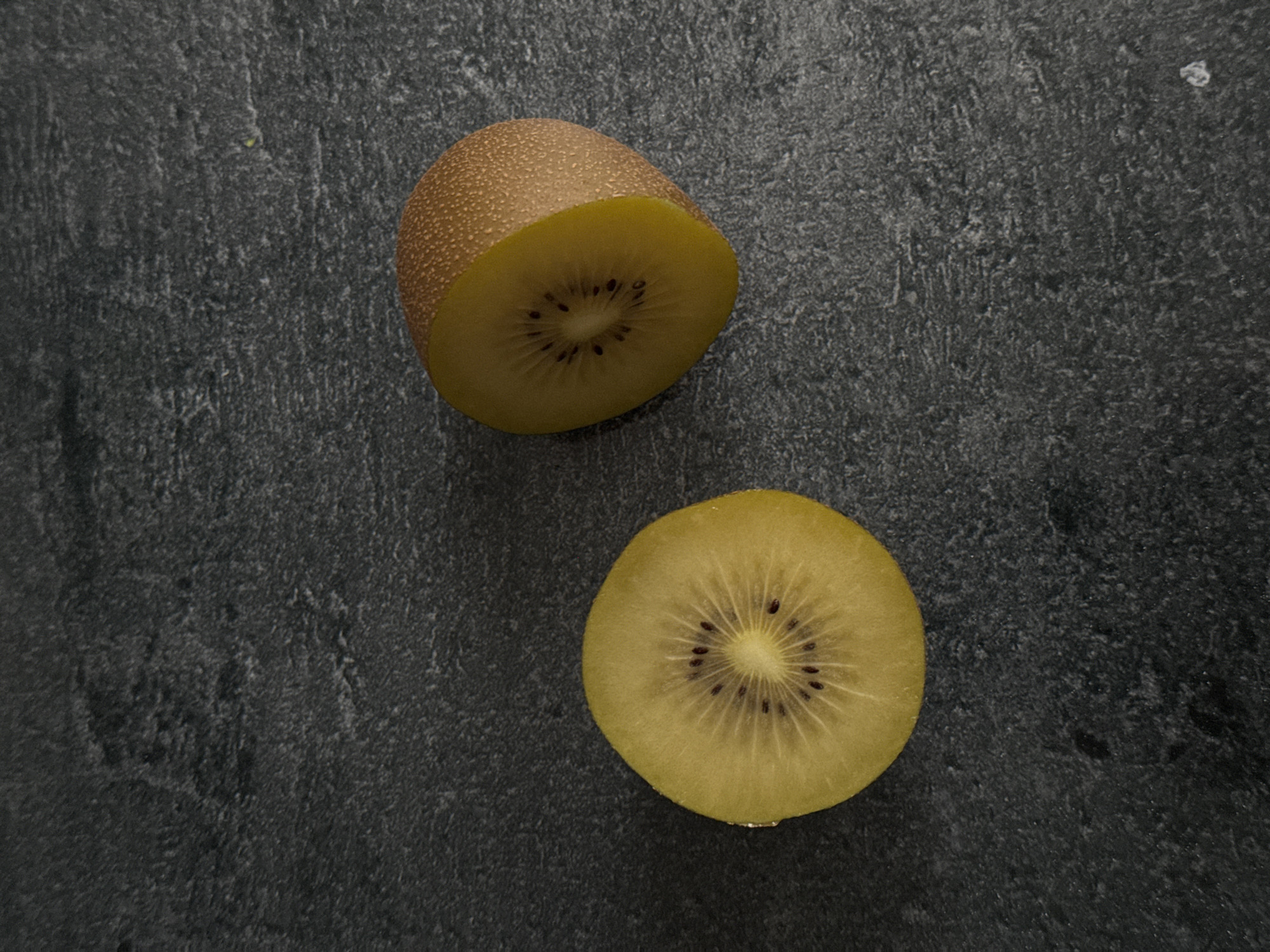 This is a high-angle, medium shot of two halves of a golden kiwi fruit, presented on a dark, textured surface. The left half is a cross-section, revealing the bright yellow flesh and the characteristic pattern of small, dark seeds arranged around a central core. The right half is a more complete portion of the fruit, showing its fuzzy, light brown skin and a portion of the yellow flesh. The dark surface appears to be a stone or concrete countertop, with subtle variations in tone and texture that create a natural, earthy backdrop. The lighting is dim, casting soft shadows that add depth and dimension to the scene. There are no people or any indication of a specific location in Siegburg, Germany, present in the image.