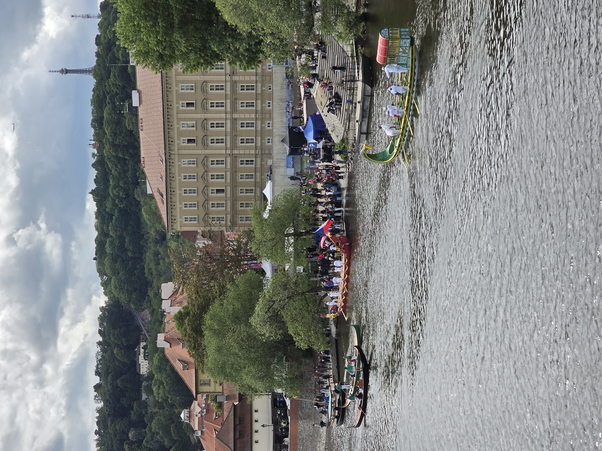 The photograph captures a vibrant daytime scene along a river, bustling with activity. On the river, several long, narrow boats are prominently featured. One boat, painted green and yellow with a decorative bow, carries multiple individuals dressed in white garments, possibly preparing for a race or procession. Another boat, red and yellow, also has people onboard and flies a flag with horizontal blue, white, and red stripes, indicative of the Czech Republic. A third, darker boat is partially visible nearer the bank.
The river banks are lined with numerous spectators, gathered to watch the event. Tents and temporary structures are set up along the bank, suggesting a public gathering or festival. A large, multi-story building with numerous windows and a tiled roof stands on one bank, complementing the traditional architecture seen further along the river with red-tiled roofs. Lush green trees line the river and cover a steep hill in the background.
The sky is partly cloudy, suggesting a bright but possibly overcast day. In the distance, atop the tree-covered hill, a distinctive, tall steel observation tower, reminiscent of the Petřín Lookout Tower in Prague, and a smaller spire are visible. The overall scene suggests a festive rowing event, parade, or cultural celebration taking place, likely in Prague, Czech Republic, given the architectural cues, the tower, and the flag. No discernible text is visible in the image.
