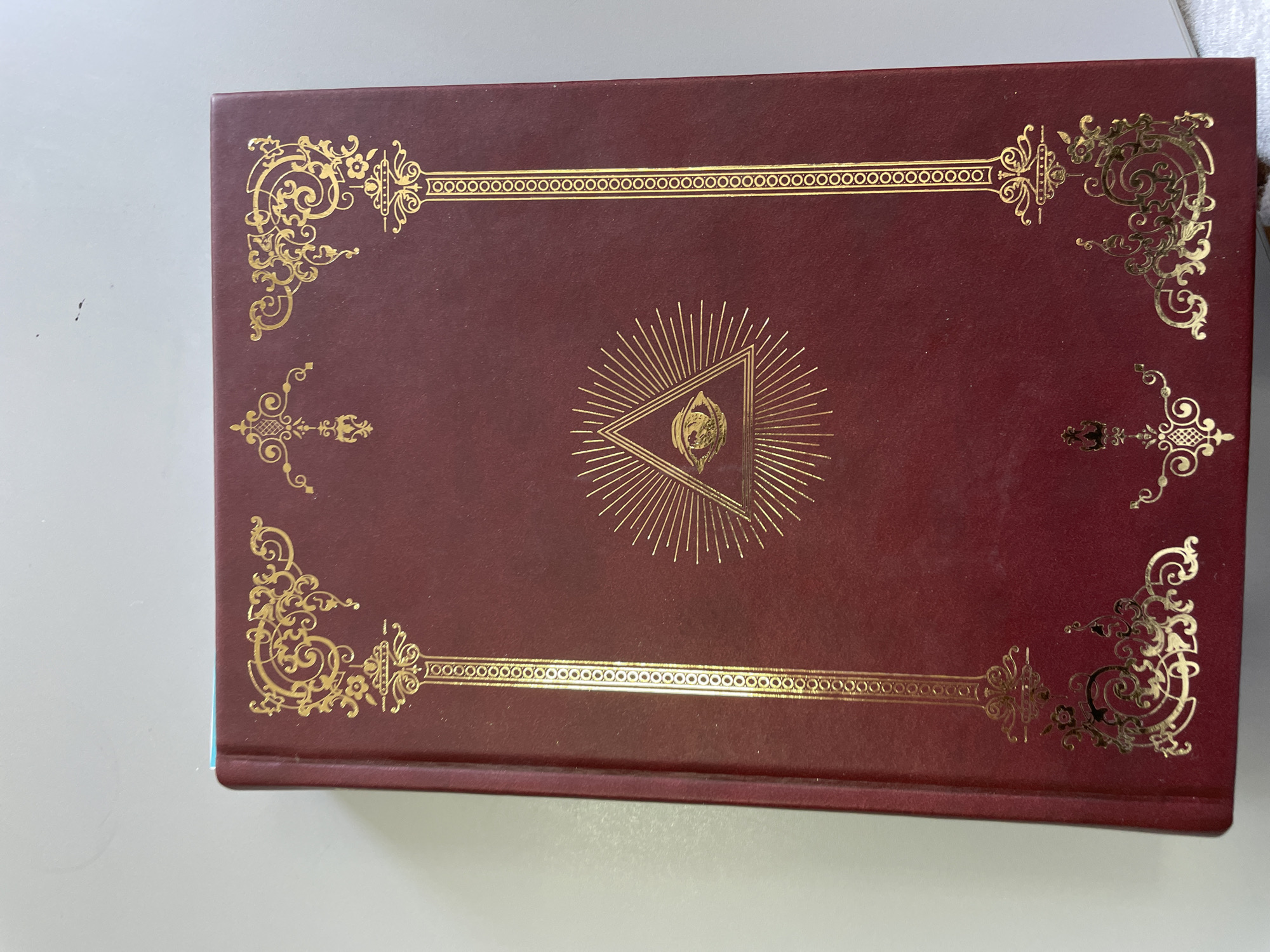 A close-up shot of a dark red book with gold embellishments. The embellishments include elaborate corner designs, horizontal line borders near the top and bottom edges, and a centered design consisting of a triangle with a circular image inside surrounded by radiating lines. The book is resting on a light-colored surface. There are no people visible. The location is unknown, and there is no indication that the image was taken in Zlatograd, Bulgaria.