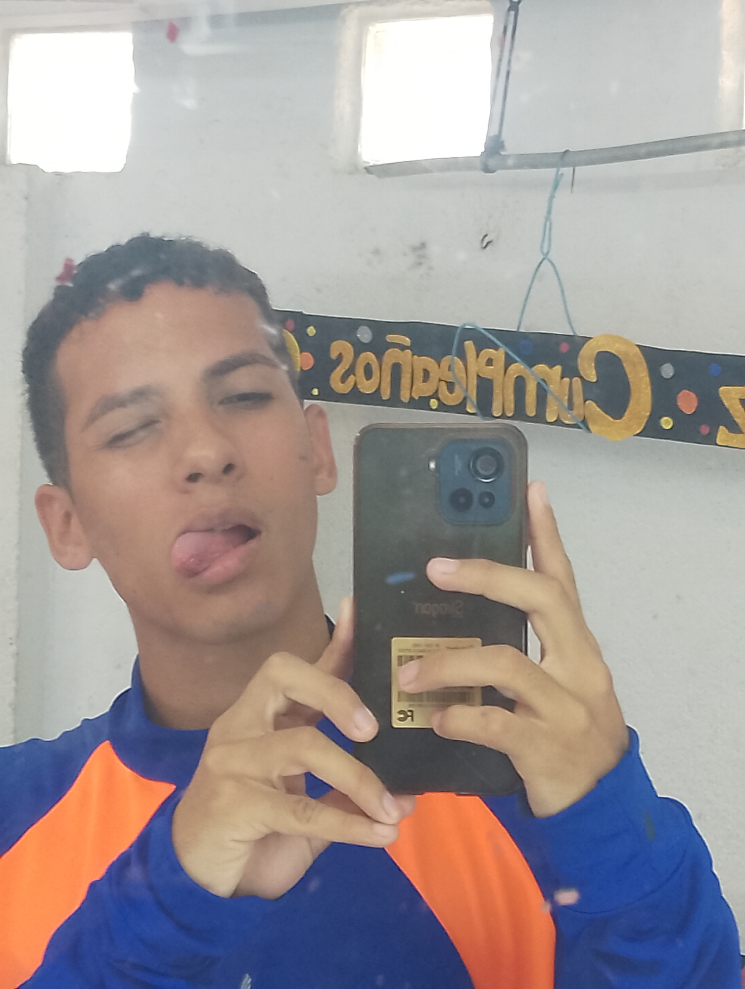 A young adult male, appearing to be in his late teens or early twenties, is depicted in a mirror selfie taken indoors in Porlamar, Venezuela. He has short, dark, curly hair and is wearing a royal blue long-sleeved shirt with an orange panel on the wearer's left side. His eyes are partially closed, and his tongue is extended to the viewer's left. He is holding a dark-cased smartphone with multiple rear cameras, consistent with an iPhone Pro or Pro Max model, in both hands. A light-colored rectangular label with reversed text, including "dopolg" and "FC," is affixed to the back of the phone. The background, reflected in the mirror, consists of a light-colored wall with two rectangular, unglazed window openings in the upper portion, allowing bright ambient light to enter. A light-colored pipe or rod runs horizontally across the upper wall, with a light blue string tied to it, which is used to hang a dark banner featuring gold letters and colorful dots. The banner's text, also reversed, clearly shows "20" and "Cumpleaños" (Spanish for "Birthday").