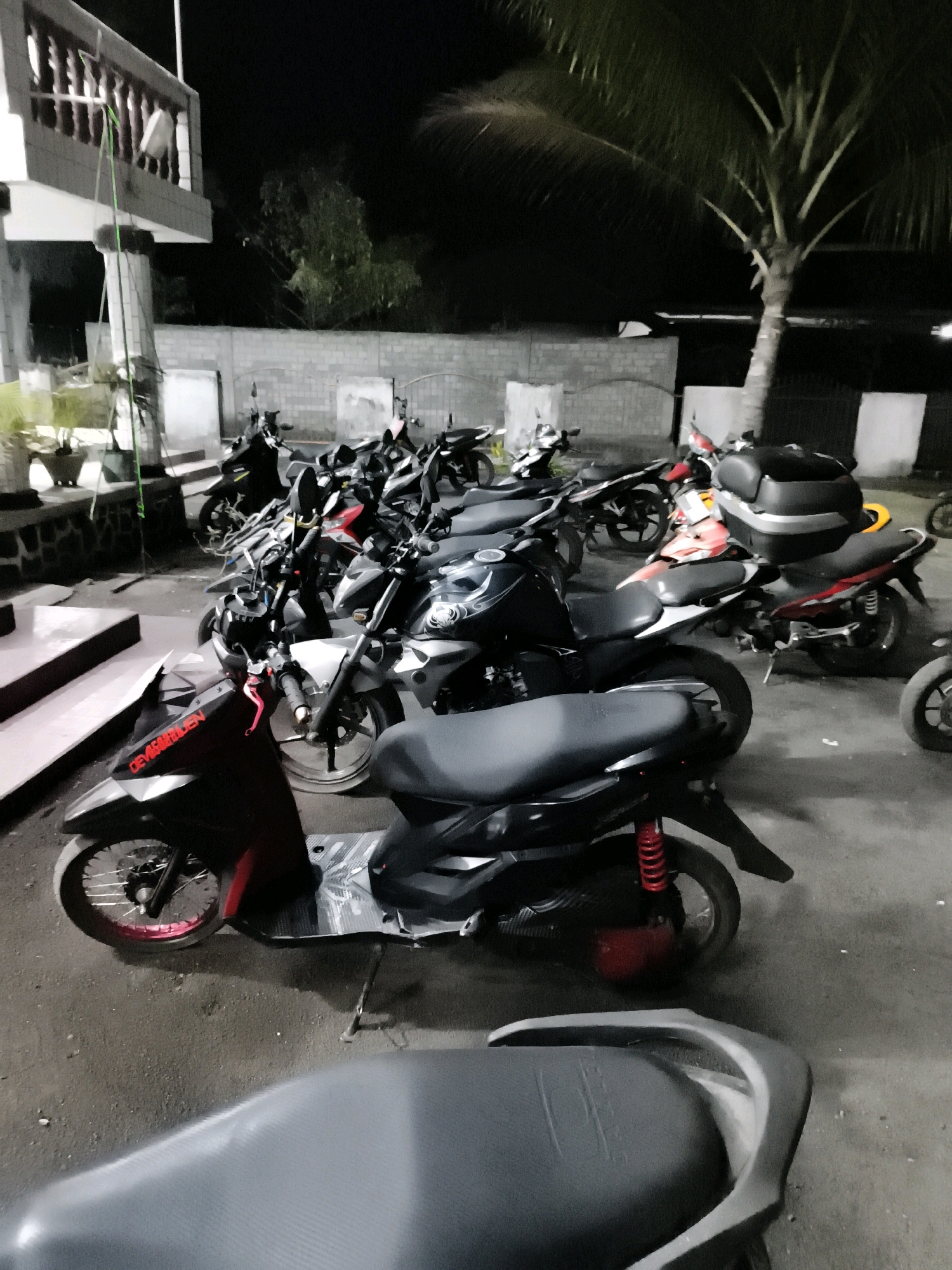 The image depicts a night scene in Tombatu Dua, Indonesia, showcasing a row of parked motorcycles and scooters. In the foreground, a black scooter with red accents and a visible kickstand is prominently displayed. Behind it, several other motorcycles, predominantly black and red, are tightly packed. One motorcycle in the middle ground features a black storage box on its rear. The background includes a building with a balcony on the left and a palm tree on the right, suggesting an outdoor area, possibly a parking lot or a street. The lighting appears to be artificial, casting shadows on the ground and highlighting the vehicles. No people are visible in the frame, and the overall atmosphere is still and quiet.