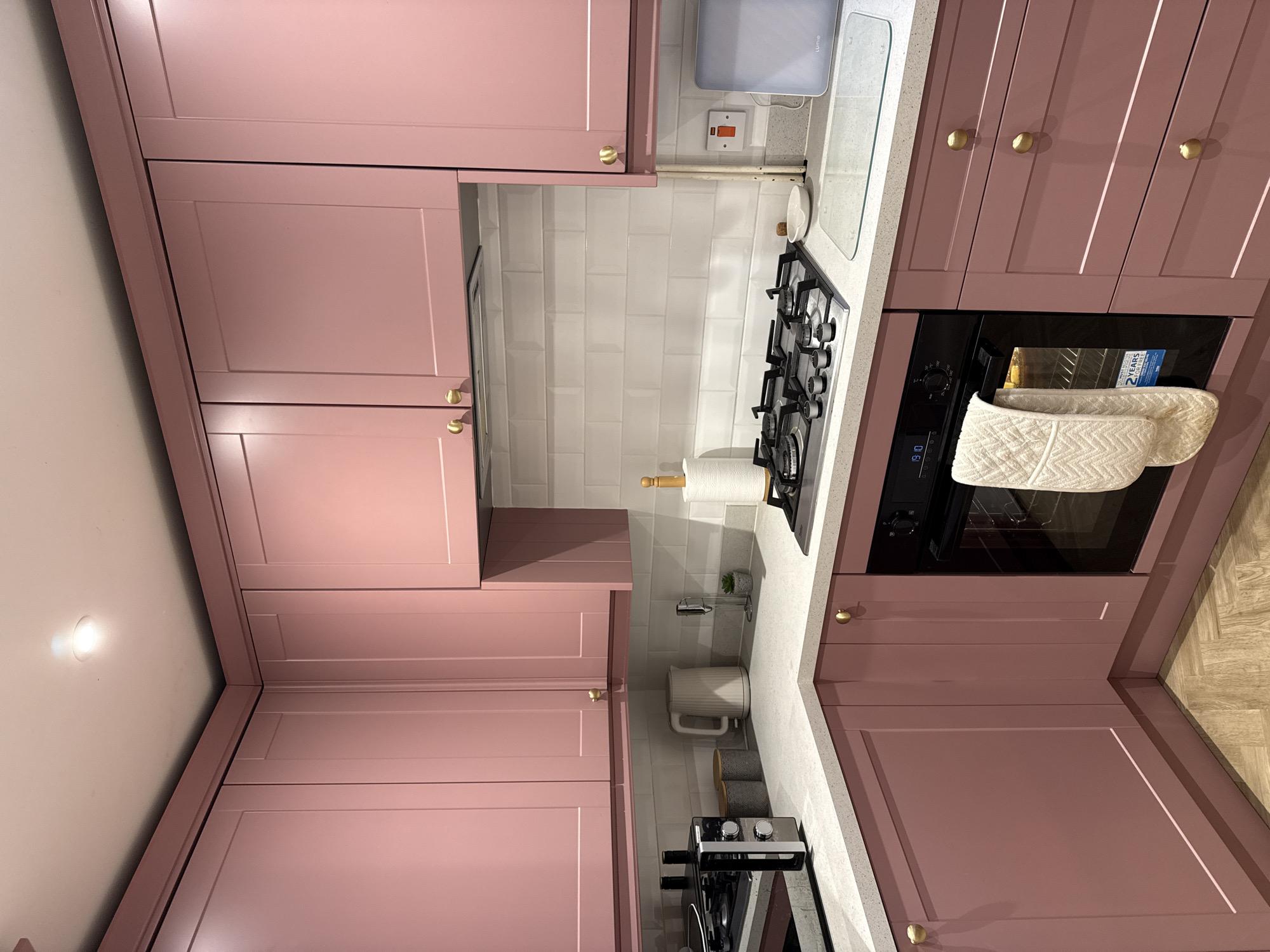 This is an interior shot of a kitchen. The cabinets are a muted pink and feature gold knobs. The kitchen countertops appear to be light grey with white, rectangular backsplash tiles. Visible kitchen appliances include a gas stovetop and a built-in oven. Miscellaneous kitchen items such as a paper towel roll, decorative plants, and mugs are on the countertop. The oven is on. The floor is partially visible in the bottom right corner, featuring a parquet pattern. The location is East Suffolk, United Kingdom.
