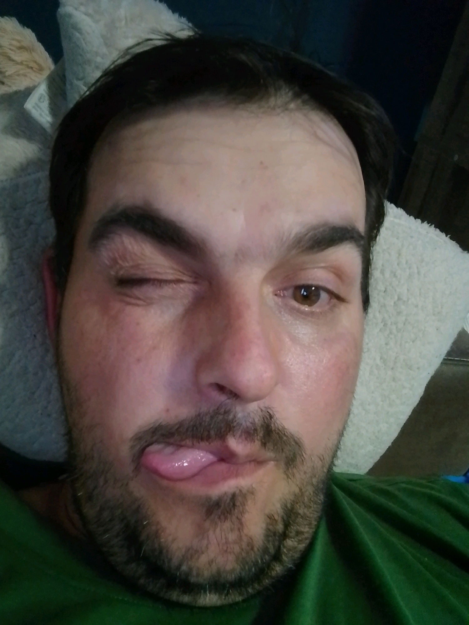 A close-up, high-angle, eye-level selfie shows a white man in his late 30s or early 40s. He has dark hair, brown eyes, and stubble. His left eye is closed in a wink, and his tongue is sticking out. He is lying down, resting his head on a beige, fluffy pillow. The background is dark and out of focus. The image was taken in Wedel, Germany.