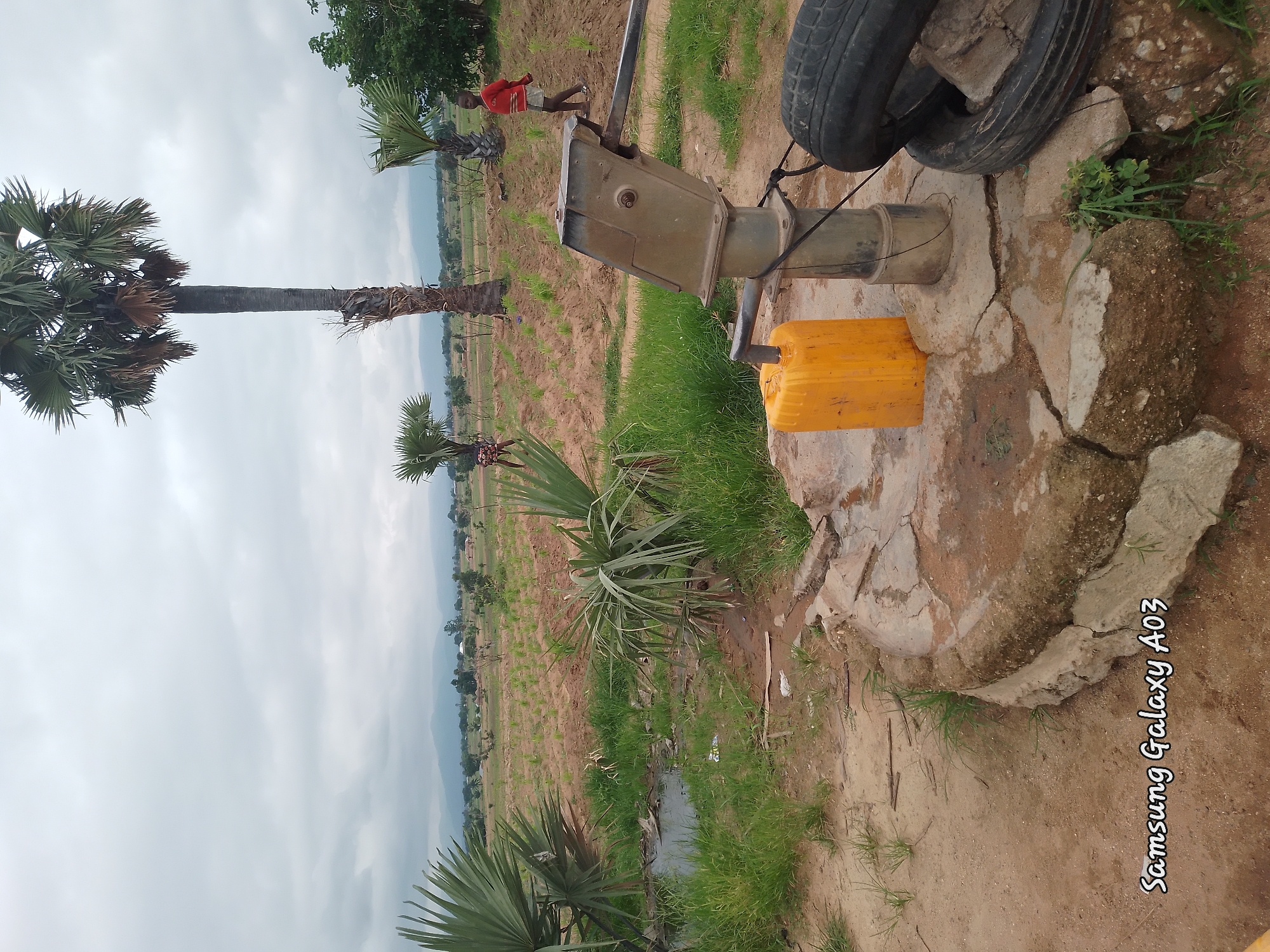The image depicts an outdoor scene at a water pump in Angwan Hausawa, Nigeria. In the foreground, a manual water pump stands on a cracked concrete base, with a yellow jerrycan positioned to its left and two car tires to its right. Behind the pump, a young boy in a red shirt and shorts stands, looking towards the right. Further in the background, palm trees and other vegetation are visible, with fields and scattered buildings suggesting a rural or semi-rural settlement. The sky is overcast. The presence of the water pump and the jerrycan indicates water collection activities. The overall impression is of a functional, utilitarian setting within a community.
