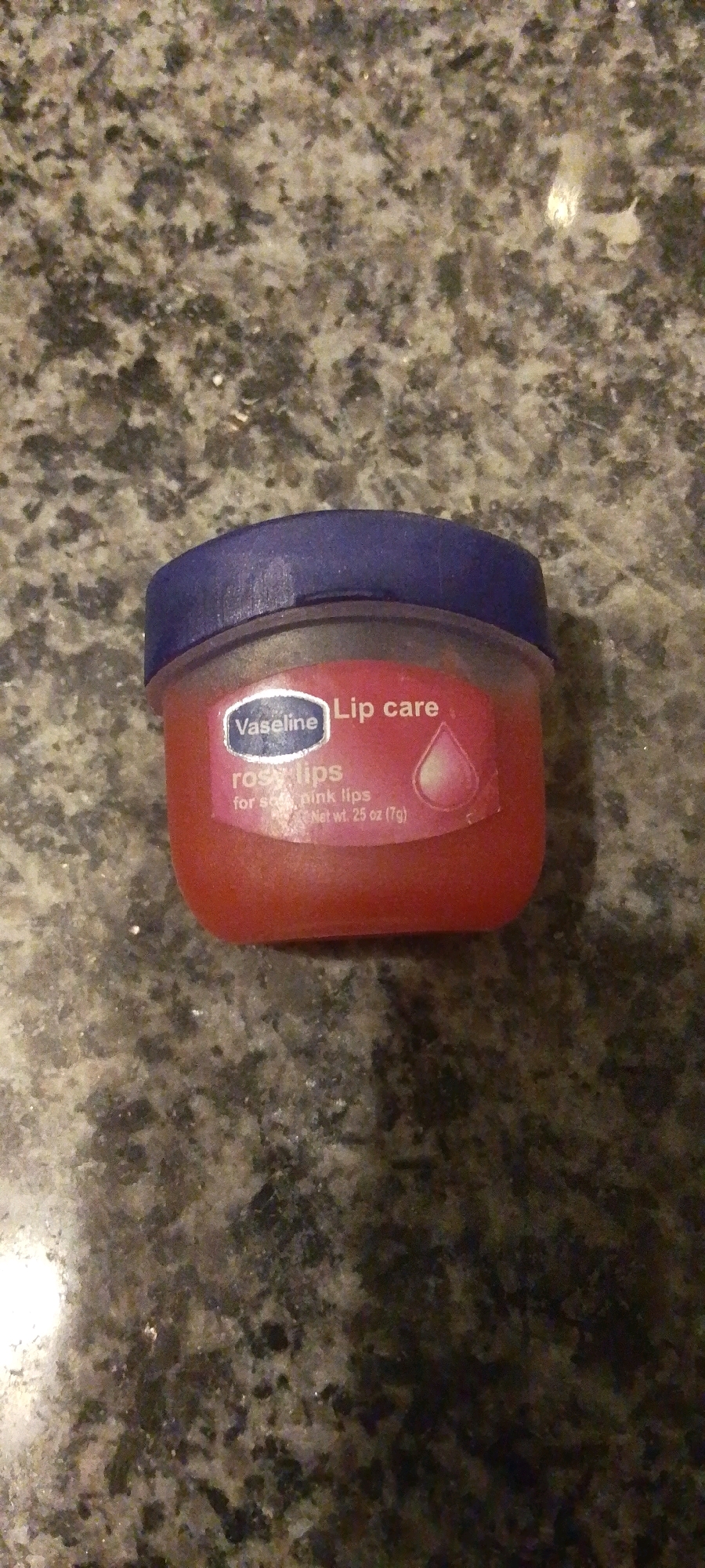 A close-up, top-down shot shows a small, round container of Vaseline Lip Care, specifically "rosy lips" lip balm, resting on a speckled granite countertop. The lip balm has a pinkish hue and a blue lid. The container's label is partially visible, displaying the Vaseline logo and text indicating "Lip care rosy lips for soft, pink lips Net wt. 25 oz (7g)". The lighting suggests an indoor setting, possibly a bathroom or vanity area. The image does not provide any information about the location in Attock, Pakistan.