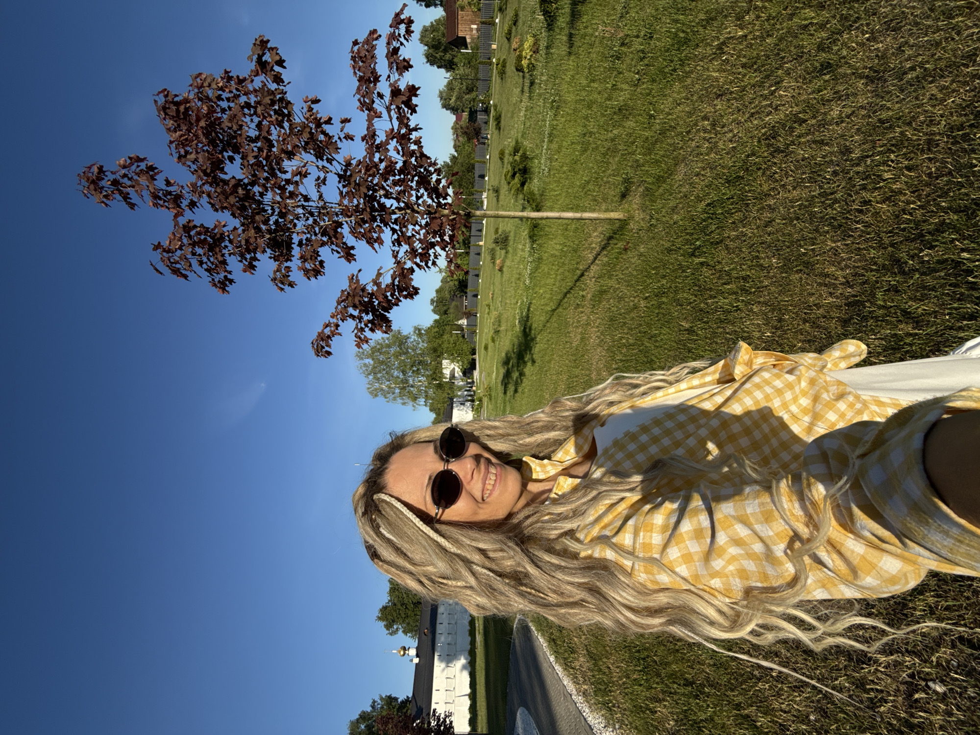 An adult female is captured in a selfie-style outdoor photograph in Dumino, Russia, under clear blue skies with bright sunlight. She is positioned in the foreground, smiling with teeth visible, and wears round dark sunglasses. Her long, wavy blonde hair is styled with numerous dreadlocks or braided extensions. She is dressed in an unbuttoned yellow and white gingham long-sleeved shirt over a white top. To her left, a light-colored paved pathway curves through a well-maintained grassy area. In the background on the left, a large, white building with a distinctive architectural style, including golden domes or cupolas on its roof, is visible, possibly a monastery or church, with an extending white wall. To the right, a young maple tree with reddish-brown leaves stands in a field of green grass dotted with smaller shrubs and ornamental plants. Further in the background, a dark fence line separates the manicured area from scattered residential-style buildings and additional trees, indicative of a village or suburban environment.