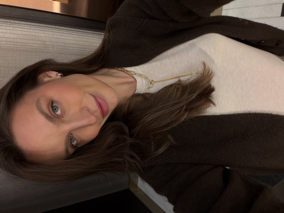A close-up, chest-up selfie depicts a female individual in an indoor setting within the city of Budva, Montenegro. The subject has long, dark brown hair, blue-green eyes, and light facial makeup including light pink lipstick and blush. She presents a slight smile and her head is tilted gently. A small, silver-toned hoop earring is visible on her left ear. She wears a gold-toned, Y-lariat style necklace featuring various oval and chain links. Her attire consists of a light-colored, possibly white, long-sleeved top, overlaid by a dark brown, textured, open-front cardigan with visible buttons along the right placket. The background reveals a vertically textured light-colored wall or panel on the left, and a darker, potentially reflective surface with a warm light reflection on the right. A small section of a light-colored tiled floor is visible at the bottom edge of the frame. The lighting is artificial and relatively dim, highlighting the subject.