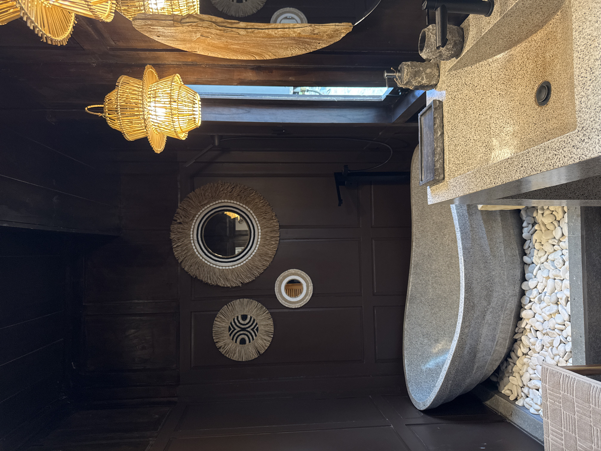 This is an interior shot of a bathroom in Ungasan, Indonesia. The walls are a dark brown, and there are three decorative circular mirrors hanging on one wall, each with a different pattern. The ceiling is also dark brown with exposed wooden beams. There is a light fixture made of woven material hanging from the ceiling. To the right is a stone sink and faucet, with a gray stone tub partially visible below. White pebbles fill the space around the tub.