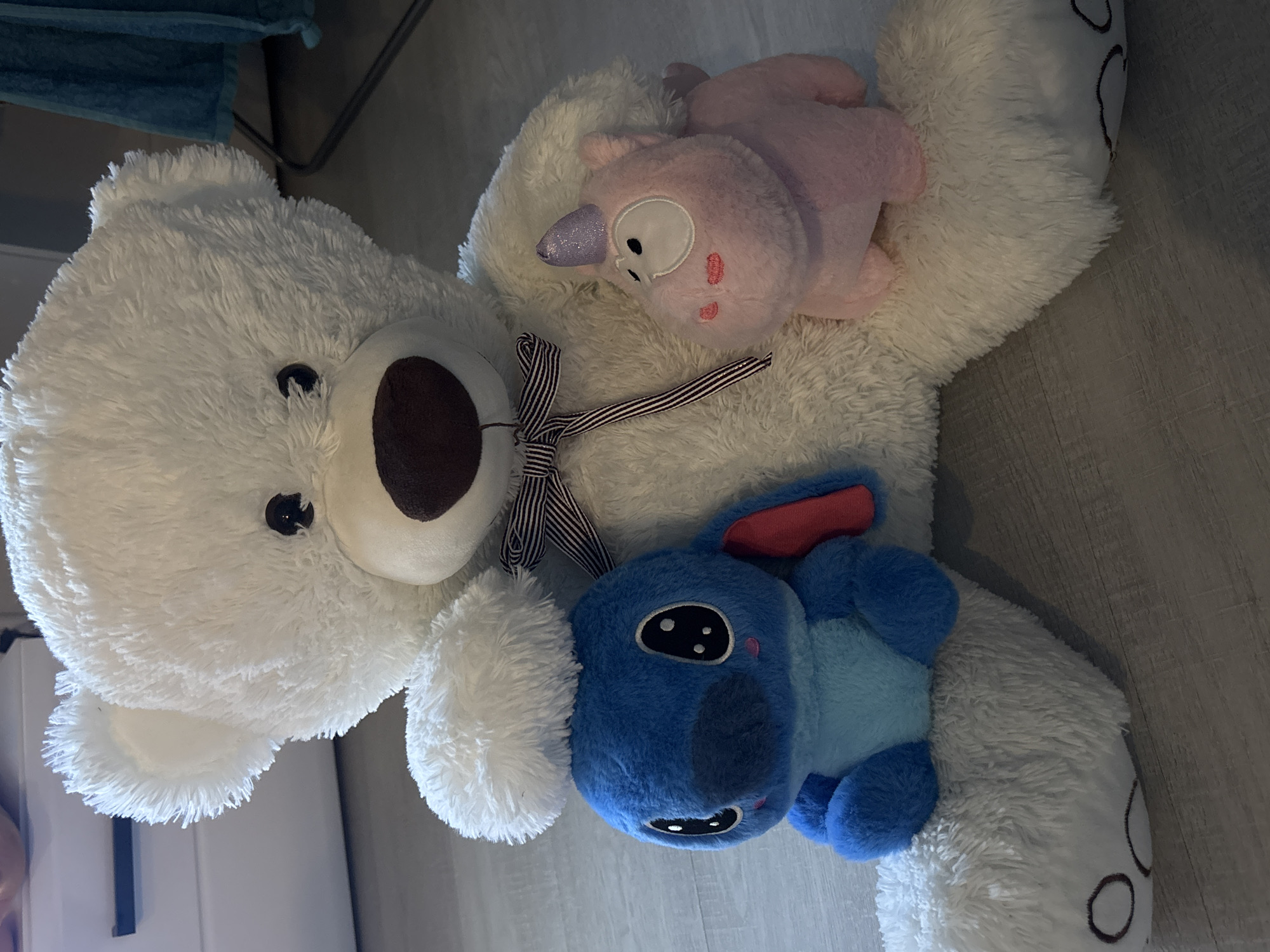 A close-up, overhead shot shows a large, white teddy bear lying on a light-colored floor. Atop the teddy bear are two smaller plush toys: a pink unicorn-like creature and a blue "Stitch" character from Disney. A striped ribbon is tied around the neck of the teddy bear. The floor appears to be laminate or vinyl. The background contains indistinct furniture and a blue cloth. The image does not contain information to determine the location in Saint Petersburg, Russia.
