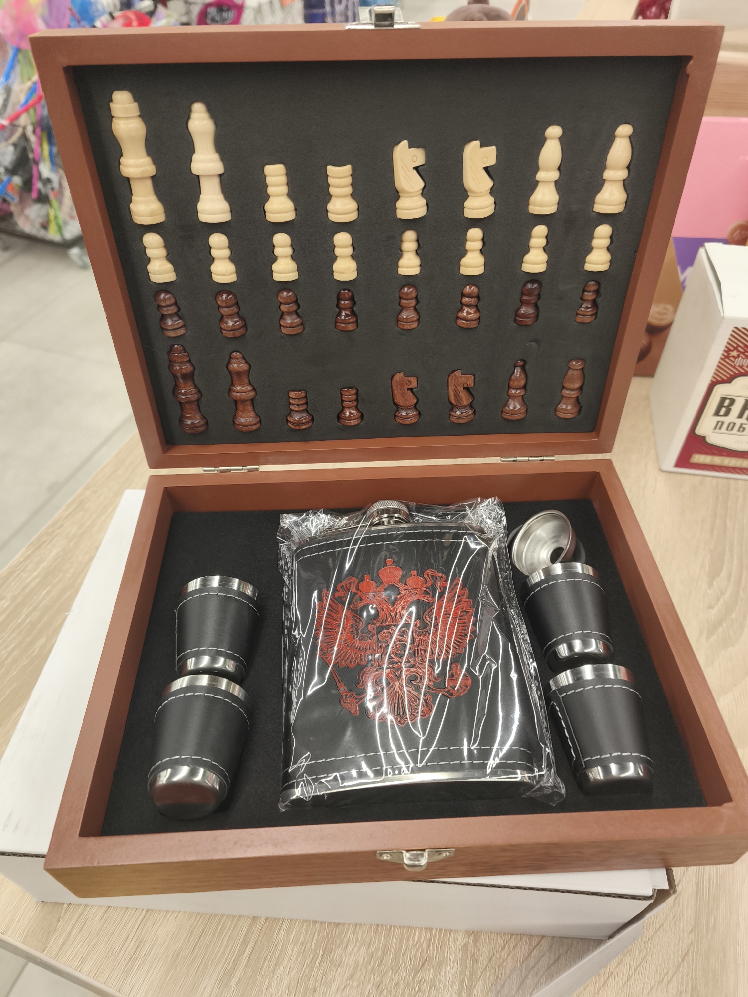 The image shows a wooden box, partially open, on a light wooden surface. Inside the box, the top compartment contains a set of wooden chess pieces, arranged in rows on a black felt background. The pieces are light wood on the left and dark wood on the right. The bottom compartment contains four small black leather cups with silver rims and stitching, a silver funnel, and a black flask wrapped in clear plastic. The flask has a red emblem on it, which appears to be a double-headed eagle. The background of the image is blurred, but suggests an indoor environment with some colorful items visible on the left and a white box with red lettering on the right. The setting is likely a retail store in Minsk, Belarus, as indicated by the presence of a box with text in Russian and Cyrillic script. There are no people or animals visible in the image.
