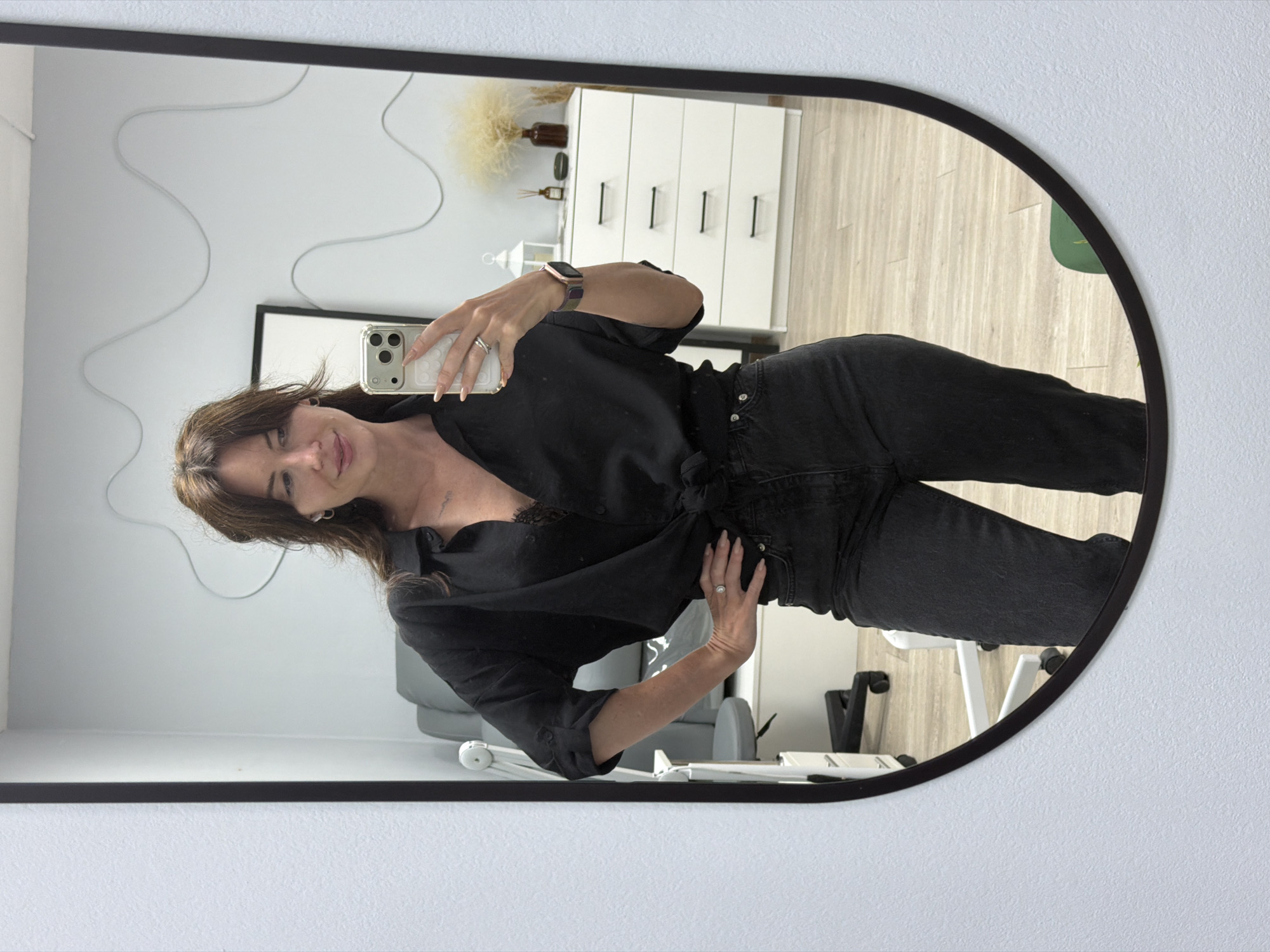 A woman is reflected in a large, oval mirror, taking a selfie with her phone. She has long brown hair, bangs, and is wearing a black, loose-fitting button-down shirt tied at the waist, revealing a black lace bralette underneath. Her pants are dark wash denim. She is wearing rings on her fingers and a smartwatch on her wrist.
The background, as seen in the mirror, appears to be a salon or treatment room. To the left, a grey chair with wheels is partially visible. Further back, a white dresser with black handles stands against a light blue wall. Decorative elements include a framed picture on the wall, a white lantern, a small bottle, and a vase with dried reeds. The floor is light wood.
The overall lighting is bright and even, suggesting artificial light. The environment is clean and organized. The specific location within the city of Perm, Russia, cannot be determined from the visual information provided.