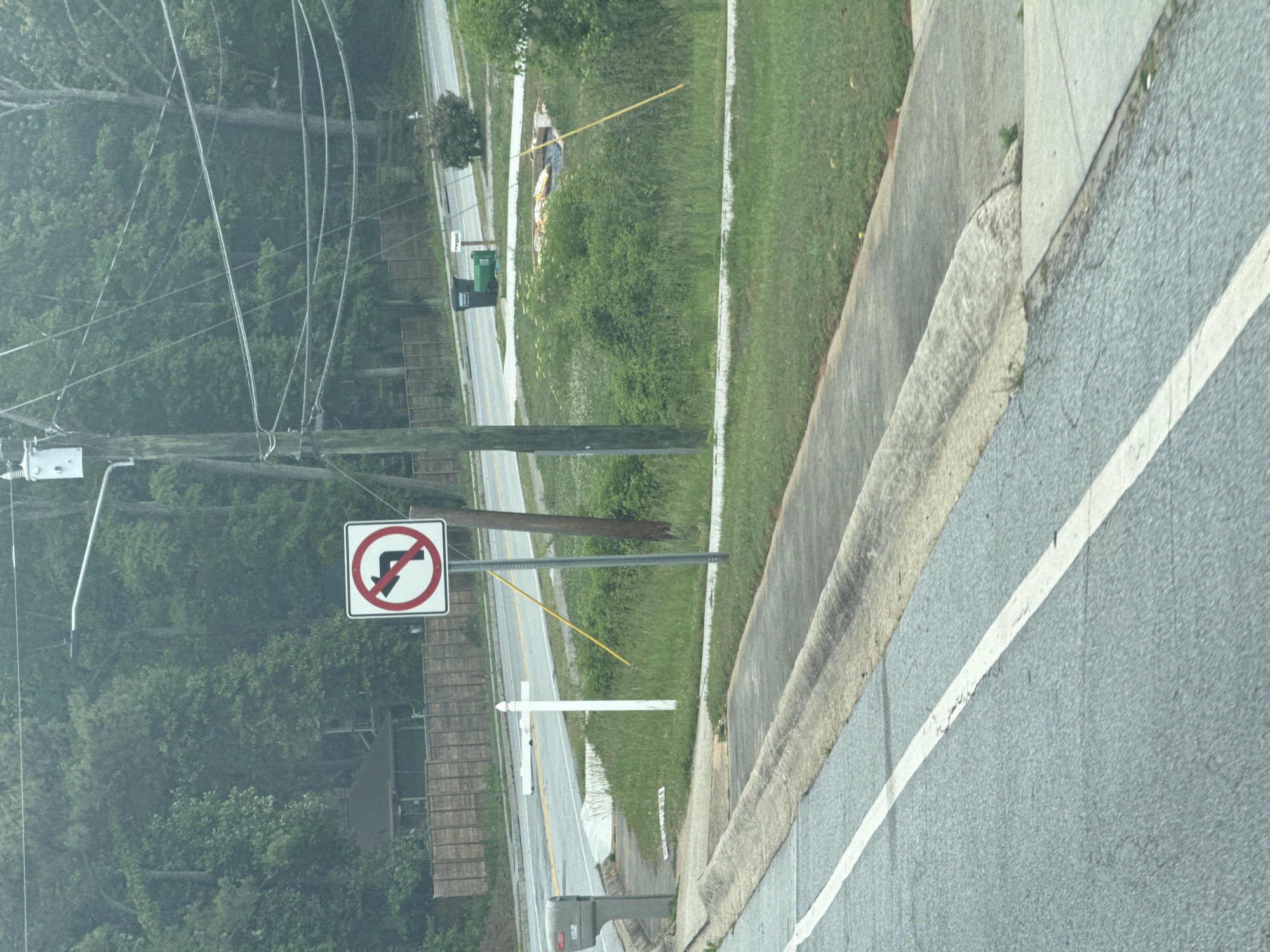 The image shows a "no left turn" traffic sign next to a road in Mountain Park, United States of America. The sign is red and white, mounted on a metal pole. A concrete curb is visible on the right side of the road, and green grass and trees are in the background. Power lines are strung overhead. A white cross is planted in the grass.