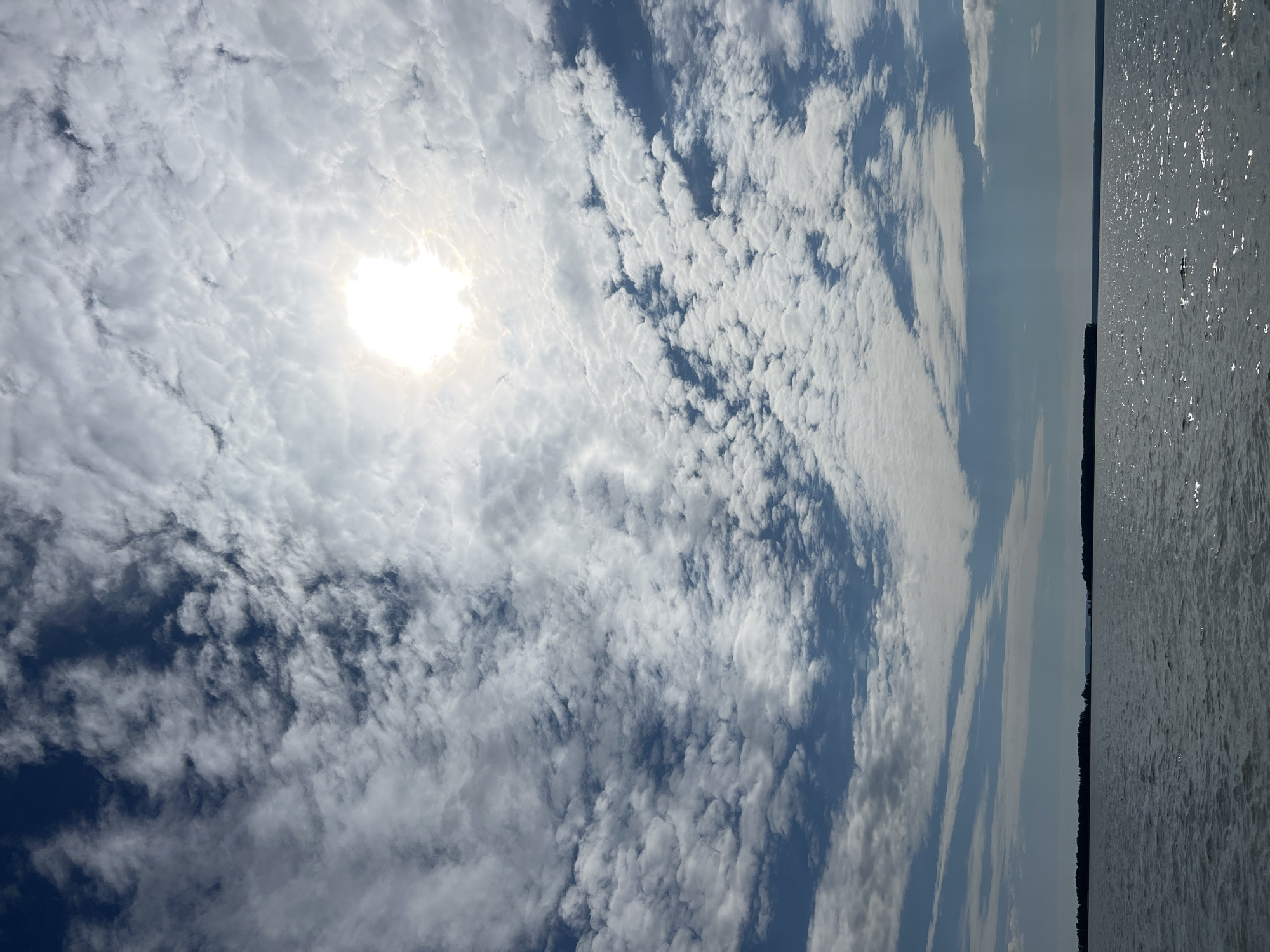 The image depicts a vast expanse of water under a cloudy sky. The sun is visible through a break in the clouds, casting a bright glow. The water appears calm with ripples on its surface. In the distance, a dark silhouette of land with trees can be seen along the horizon. The sky is a mix of blue and white, with scattered clouds of various sizes and densities. The overall scene is tranquil and natural, evoking a sense of open space and natural beauty. The location is described as Novopichugovo, Russia, implying a scenic natural setting possibly near a body of water such as a lake or river. There are no people or specific man-made structures visible in the image, focusing solely on the natural landscape.