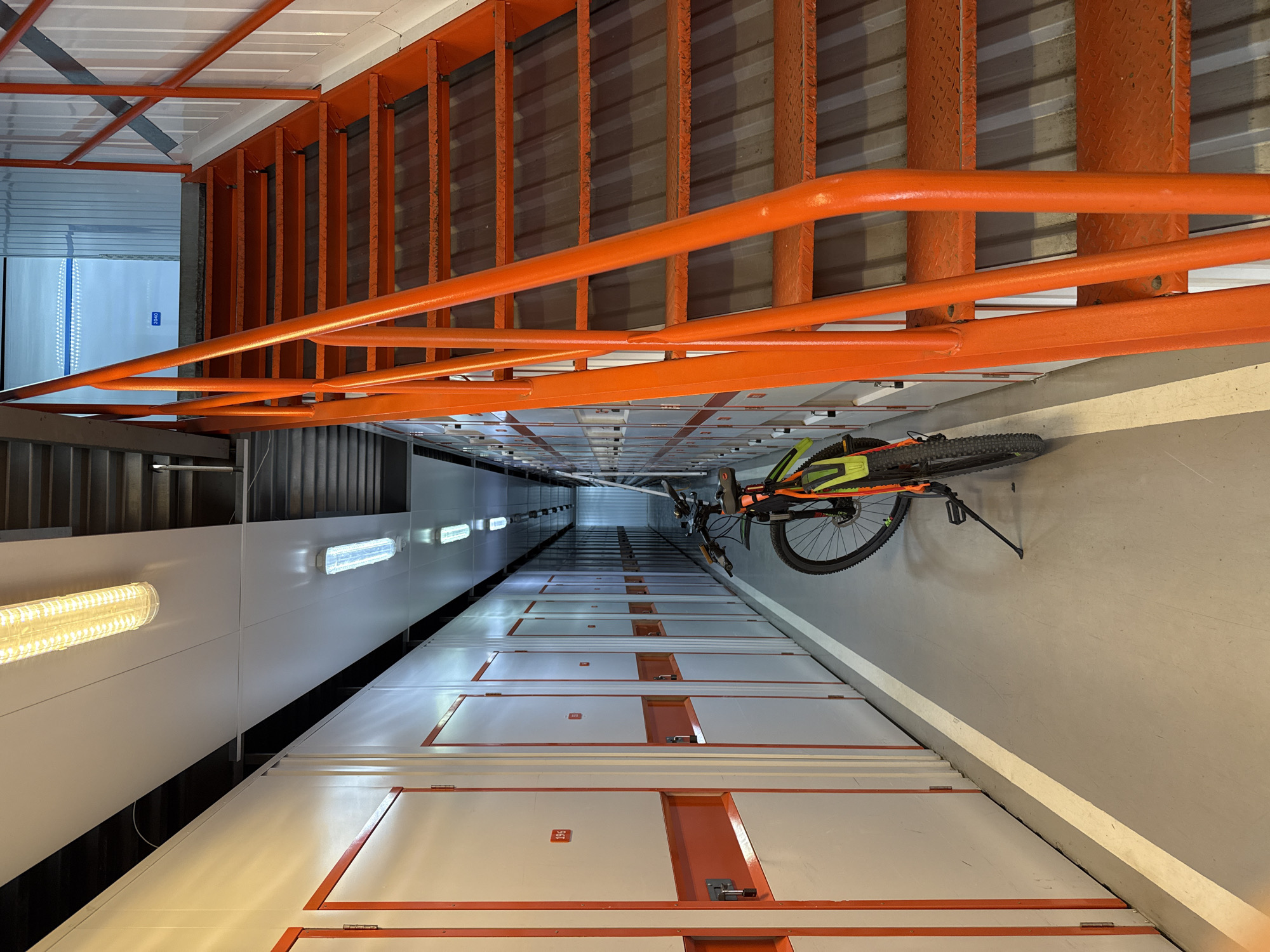 The image shows the interior of a storage facility, featuring rows of white storage units with orange trim. A green and orange mountain bike is leaning against the wall on the right side. Above the bike, an orange metal ladder is mounted to the wall. Fluorescent lights are visible along the left wall. The location details are not evident in the image; therefore, identifying the city of Moscow, Russia, is not possible based on the visual information.