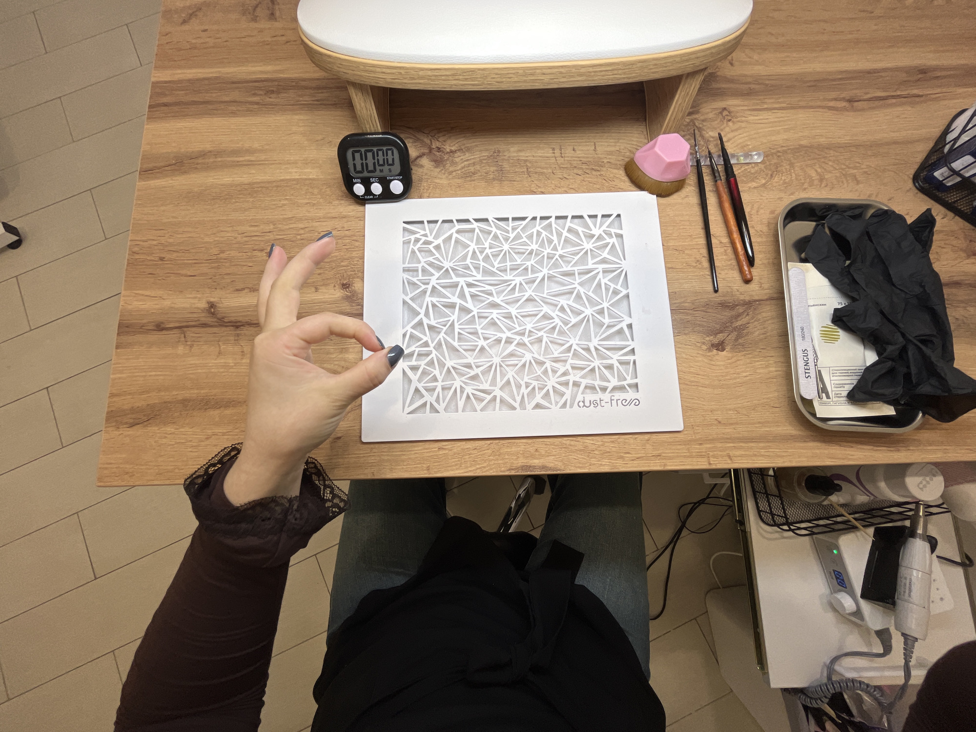 The scene depicts an overhead view of a nail care workstation located in the city of Neftekamsk, Russia. A person is seated at a light brown wooden table, with their right arm and hand visible in the upper-left quadrant. The individual wears a dark brown long-sleeved top with a lace-trimmed cuff. Their right hand, with grey/blue polished fingernails, is positioned above a white square-shaped object, with the thumb and index finger touching. The individual's legs, clad in light blue jeans and a dark garment, are partially visible beneath the table.
On the wooden tabletop, a white rectangular object, approximately 25cm x 25cm, is centered. This object features the text "dust free" and a cut-out geometric pattern composed of multiple interlocking triangles. To its right, a black digital timer displays "0000" and has buttons labeled "CLEAN," "SET," "MIN," and "SEC." A white cushioned armrest with a light wooden frame is visible on the far right. Below this armrest, a pink faceted brush with tan bristles, three slender nail art brushes with varying handle colors (black, brown, dark red), and a clear strip containing small decorative elements are arranged. In the lower-left corner of the table, a silver metal tray holds a white nail file marked "STENGUS 180/240," a folded paper with green graphics, and several crumpled black nitrile gloves.
Under the left side of the table, a white electronic nail drill machine with a silver dial, a small illuminated display showing "00," and a green indicator light is visible. A white nail handpiece is connected to it by a coiled grey cable. Multiple black power cords and cables are also present, along with a black wire mesh basket containing white cylindrical bottles. The floor beneath the table is covered with light-colored rectangular tiles.