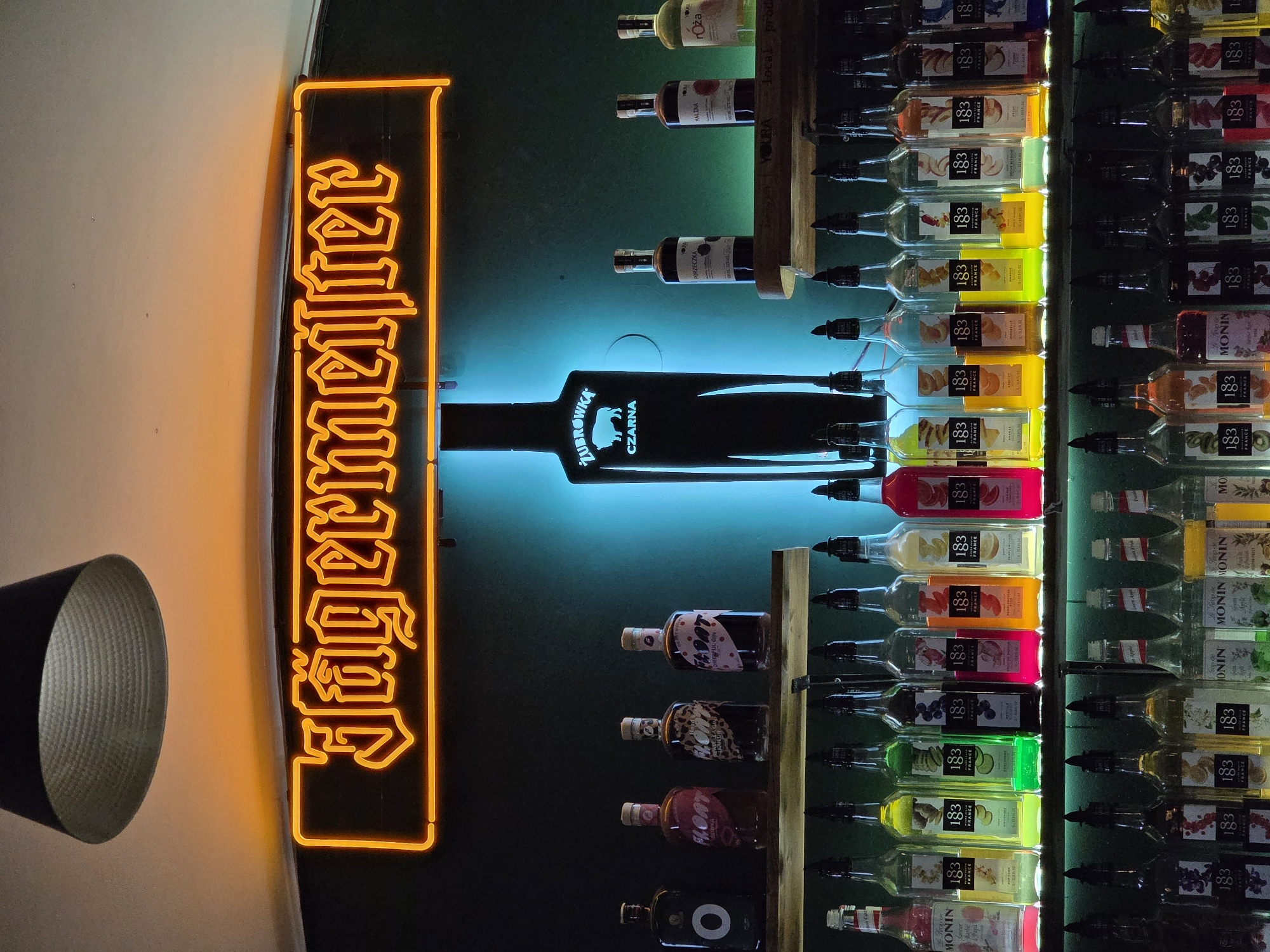 The image depicts the interior of a bar or establishment in Krakow, Poland, featuring a prominent neon sign that reads "Jägermeister" in orange lettering, set against a dark green wall. Below the sign, a large, black, stylized bottle shape is illuminated from within with blue light; this bottle shape features the Zubrowka logo with a bison and the word "CZARNA."
On a shelf directly below the neon sign and bottle, there are several bottles of spirits, including four bottles of "FLOAT" brand alcohol.
Further down, two more shelves are filled with an array of colorful bottles, primarily syrups. The top shelf of syrups displays bottles from the "1883 France" brand, with various fruit flavors such as raspberry, lemon, kiwi, blueberry, strawberry, peach, plum, apricot, and mango. There are also some bottles from the "Monin" brand visible on this shelf, including "Le Sirop de Monin" with "Charlie & The Chocolate Factory" branding and "Le Sirop de Monin Passion Fruit."
The bottom shelf also contains "1883 France" syrup bottles, with flavors like cherry, apricot, strawberry, blueberry, and possibly others. Additionally, there are more "Monin" syrups, including "Green Apple," "Melon," and "Peach."
The lighting in the scene is dim, with the neon sign, the illuminated Zubrowka bottle, and the colored syrups providing the primary light sources. A dark, textured lampshade is visible in the upper left corner of the image. The overall impression is of a well-stocked bar, likely a popular spot for drinks.