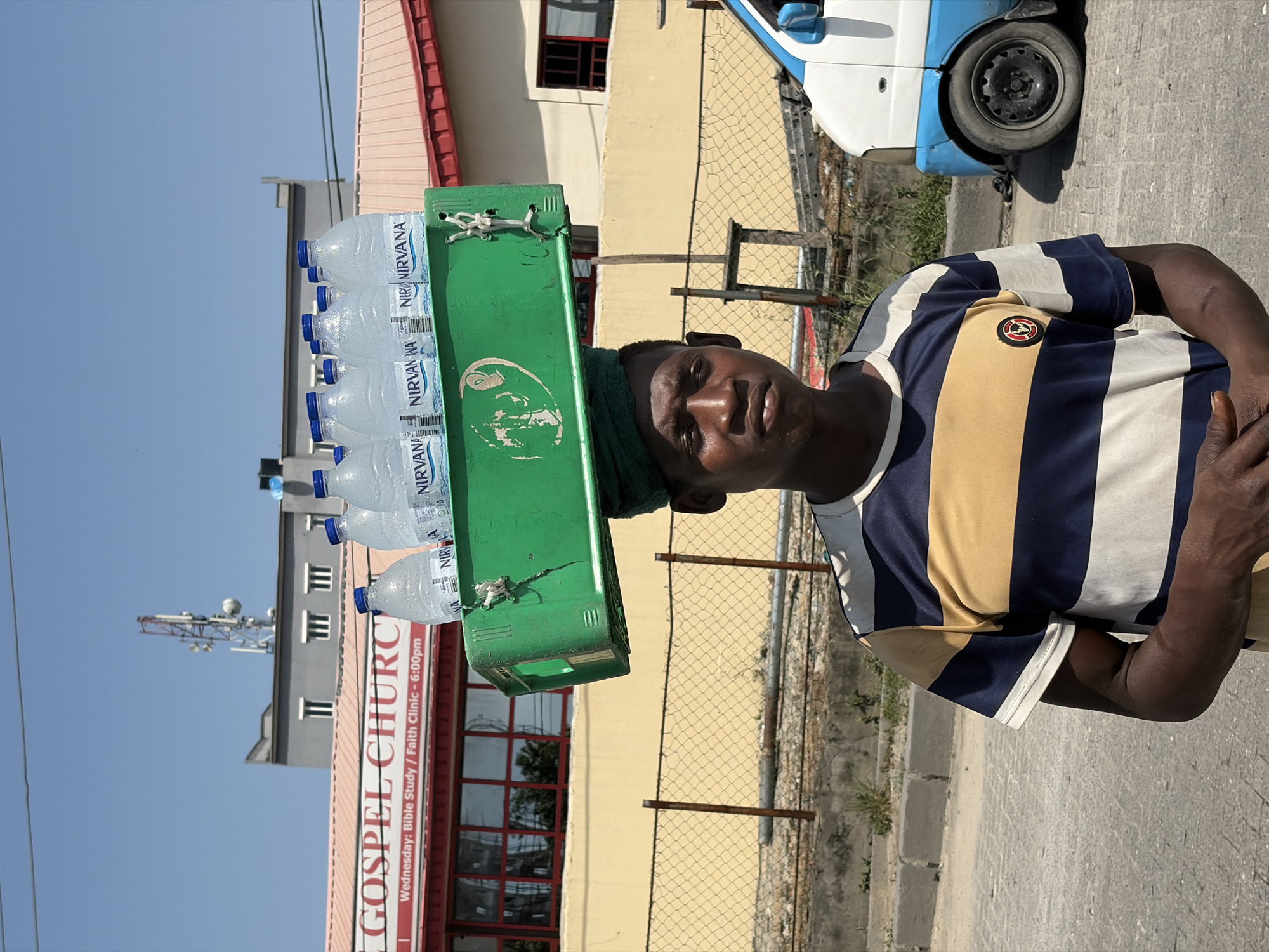 A street vendor carries a green crate filled with plastic water bottles on his head in Igboefon, Nigeria. The man, with dark skin and a neutral expression, looks directly at the camera. He wears a short-sleeved striped shirt in dark blue, tan, and white. A dark green cloth or towel is visible under the crate on his head, presumably for padding.
The plastic bottles are clear with blue caps, and the visible label on several bottles clearly reads "NIRVANA". The green crate appears well-used.
In the background, there's a paved street and a mesh fence. Various buildings are visible, including a cream-colored building with red-framed windows and a red-roofed structure. Another building in the distance, partially obscured, has the red sign for "GOSPEL CHURC" (partially cut off) with text below it stating "Wednesday: Bible Study / Faith Clinic - 6:00pm". A cell tower is visible behind this building against a clear blue sky. A white and blue tricycle vehicle, resembling a Keke Napep, is parked on the right side of the frame. The scene is brightly lit, indicating a clear, sunny daytime.