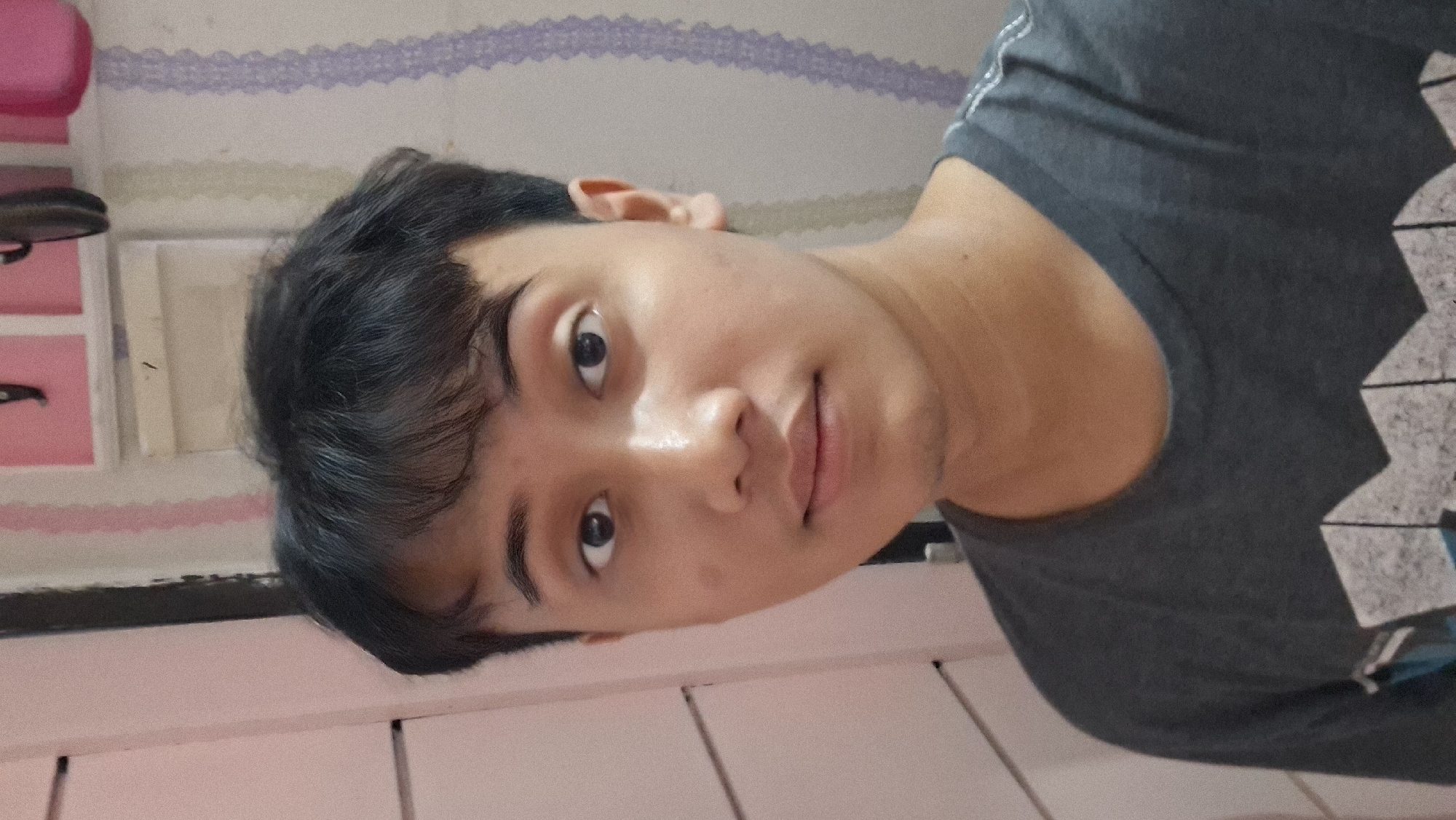 An eye-level portrait features a young person, likely male, with short, dark, slightly wavy hair and dark eyes. The individual has light to medium skin tone and a neutral, calm expression, looking directly into the camera. There is a slight sheen on their forehead and cheeks, suggesting either lighting or natural skin oils. They are wearing a dark grey t-shirt with a graphic design of angular, geometric shapes, predominantly white and blue, visible on the right side of the chest.
The background indicates an indoor setting, likely a personal room in Ciseeng, Indonesia. To the left and behind the person's head, a light-colored shelf or cabinet is visible, featuring pink storage elements or drawers, and some white rectangular objects. Above this, the wall is adorned with decorative wallpaper or border patterns; a distinct purple, lace-like horizontal pattern runs along the top, beneath which are horizontal stripes and wavy green/white patterns. The lower part of the background, below the person's shoulder, shows a light-colored surface, possibly a desk or bed frame, with vertical lines suggesting panels. The lighting is soft and even, typical of indoor conditions, with no specific time of day discernible. No visible text is present in the image.