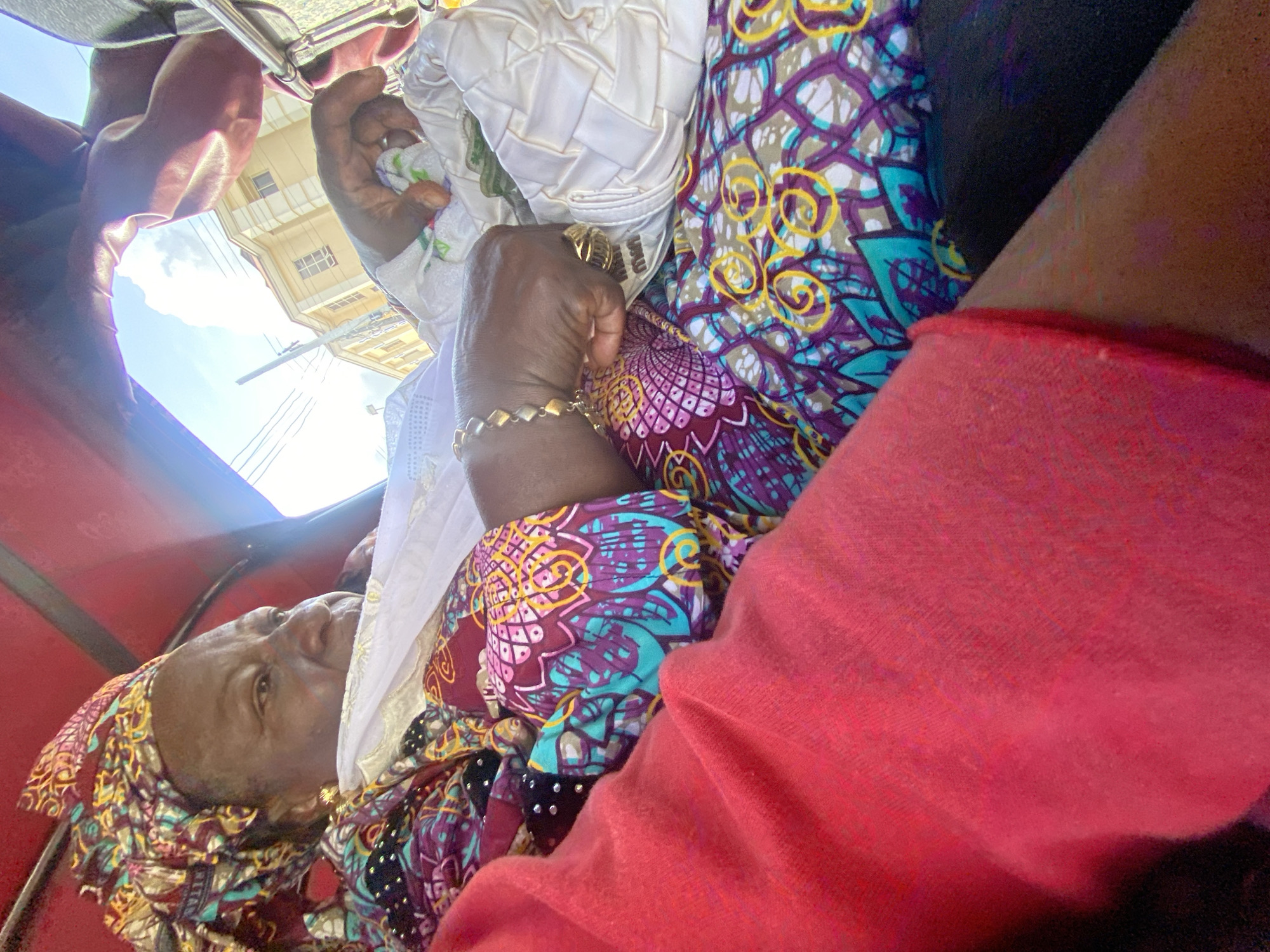 In Ado, Nigeria, an elderly Black woman, presumably within a vehicle, is captured from a low-angle, close-up perspective. She wears a patterned headscarf and a colorful top with purple, teal, and yellow motifs. Jewelry adorns her wrist and fingers. She is holding a white woven purse. Outside the vehicle, a building under a partly cloudy sky is visible. The vehicle's red interior frame surrounds the scene. A person wearing a red top is partially visible in the bottom right corner of the frame.