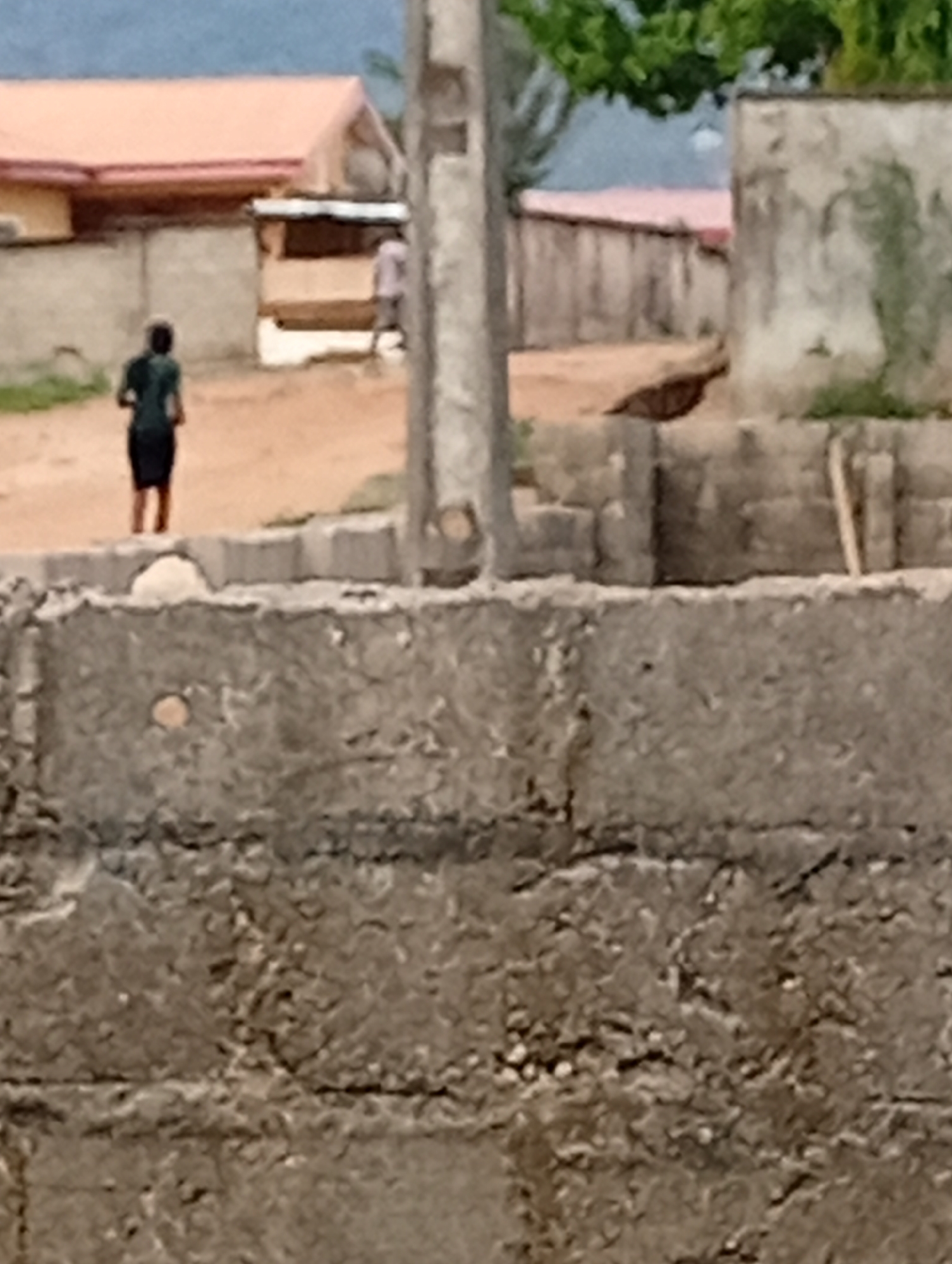 The image captures an outdoor scene, likely a residential or developing area, dominated by various concrete structures and dirt ground under daylight conditions. The foreground is occupied by a textured, grey concrete block wall, appearing rough and possibly unfinished, with some small holes or imperfections.
In the middle ground, behind this wall, a dirt path or open area extends into the background. Two human figures are visible walking on this ground. On the left-center, a person, viewed from behind, wears a dark top and a dark skirt or shorts, with bare legs. They are walking away from the camera. Further to the right and slightly behind them, another person is partially visible, mostly obscured by a structure, wearing lighter-colored clothing (possibly a light shirt).
The background features several buildings. A prominent structure on the left has a reddish-orange gabled roof and light-colored walls. Other less distinct structures and walls made of concrete blocks are also present. Some green foliage from trees or bushes can be seen at the top of the image, suggesting vegetation in the area. The overall impression is of a casual, everyday scene in a possibly warm climate. There is no visible text in the image.