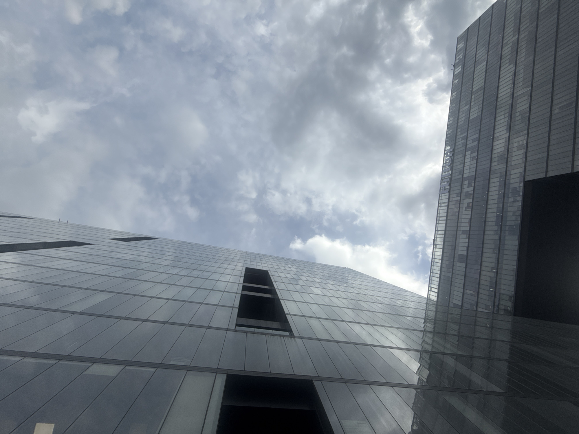 A low-angle shot showcases two modern skyscrapers against a backdrop of a cloudy sky. The building on the left is characterized by a grid-like facade of rectangular windows reflecting the sky. It appears to have some unfinished areas or voids where the windows are absent. To the right is a taller building, also with a glass facade, partially cut off by the frame. Its darker reflective surface contrasts with the lighter sky. The cloudy sky contributes to the scene's overall muted and grey tone. Given the architectural style and location, the scene likely represents a view within Mexico City, Mexico.