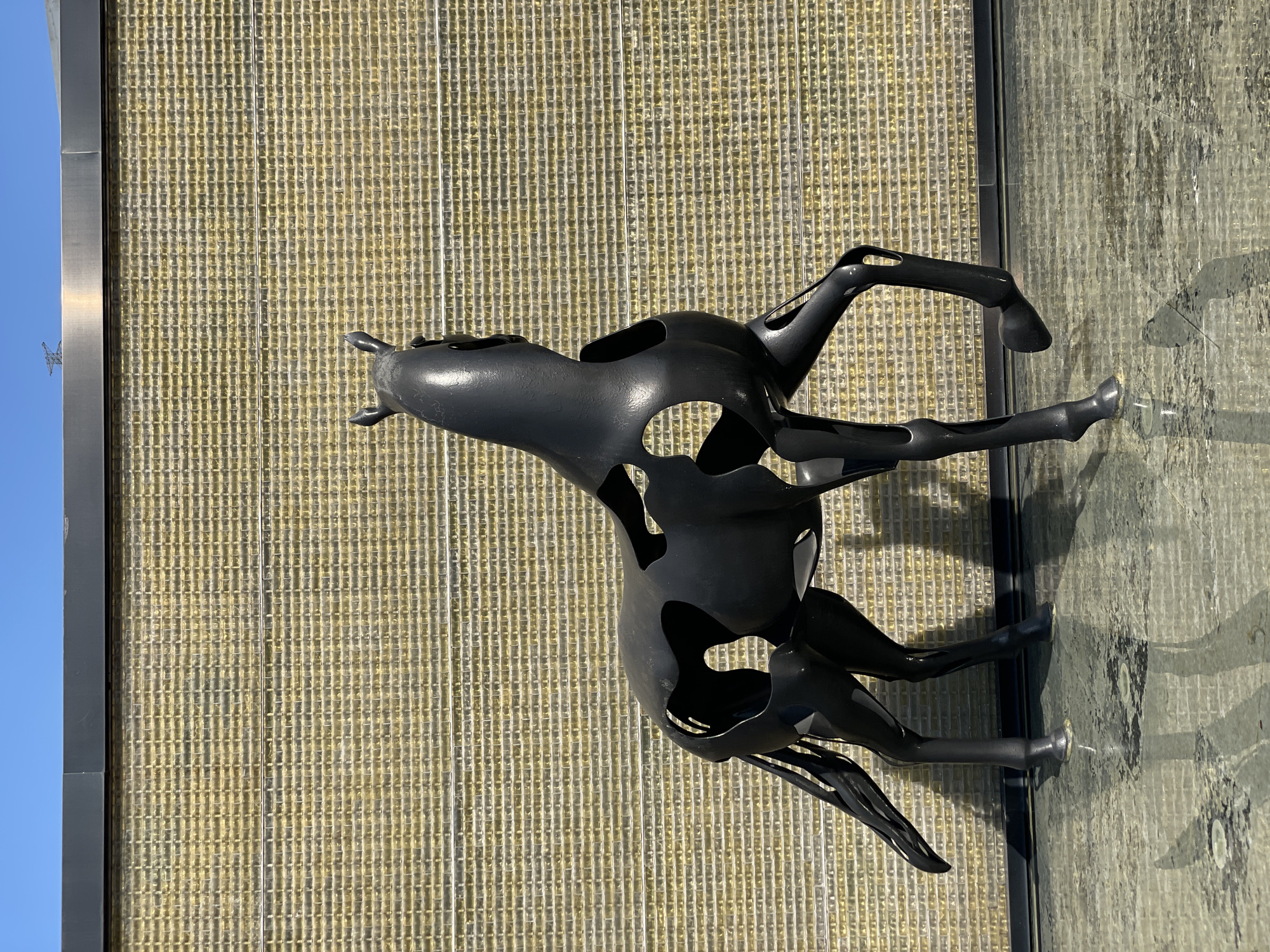 A black, hollowed-out sculpture of a horse stands on a reflective surface in front of a textured wall. The wall is made of small, rectangular, yellow-toned blocks arranged in a brick pattern. A dark grey band runs horizontally across the top of the wall. The sky above is clear and blue. The horse sculpture has large cutouts, revealing its hollow interior. Its tail is extended backward, and its legs are positioned as if in motion. The reflective surface below shows distorted reflections of the sculpture and the wall. The overall scene appears to be outdoors. The location is identified as Nan'an District, China.