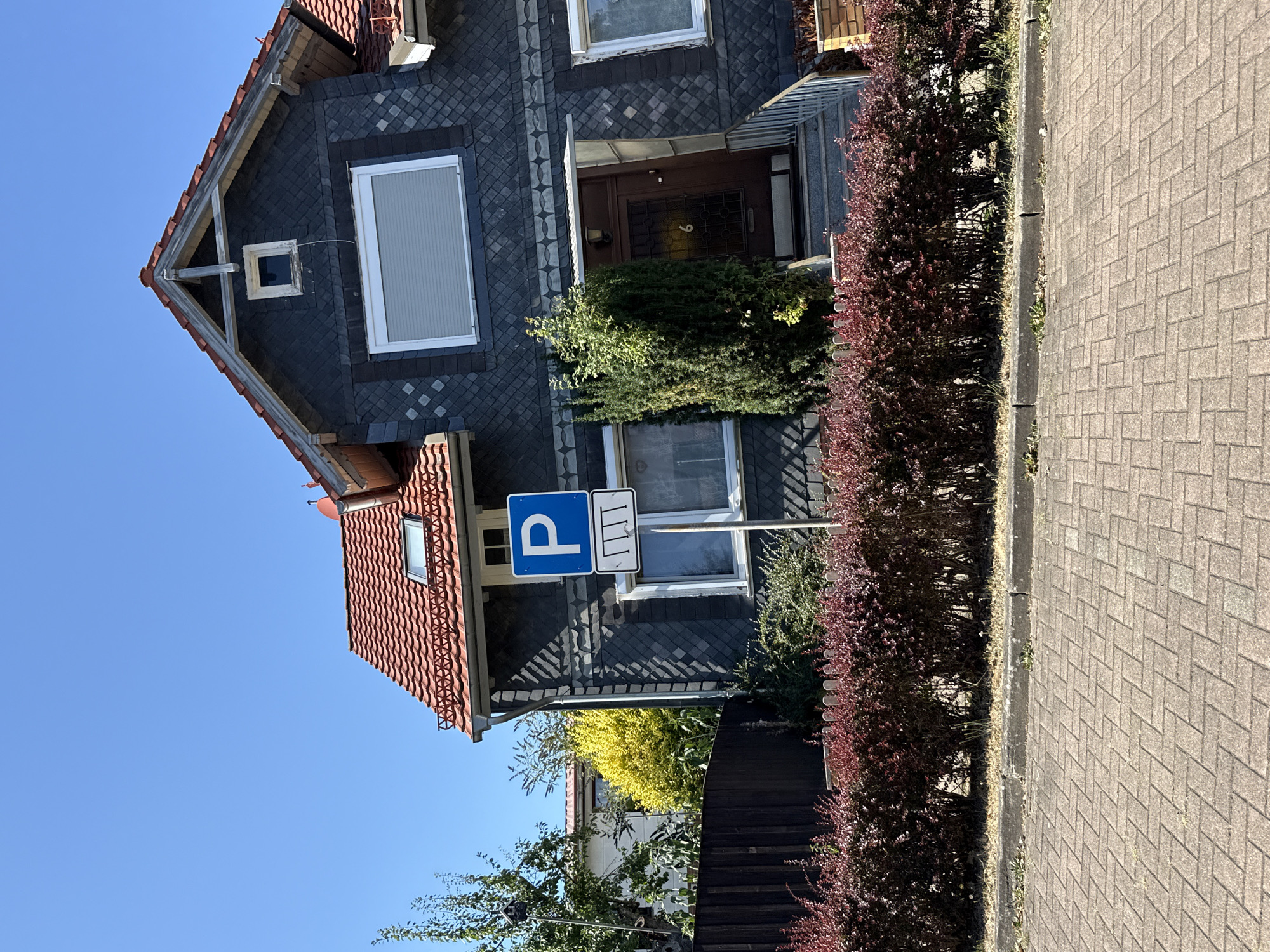 The image shows a residential building with dark grey slate siding and a red tiled roof. In front of the building, there is a paved walkway made of rectangular bricks, a row of red-leafed bushes, and a parking sign. The parking sign is blue with a white "P" and a white symbol below it depicting vertical lines. The building has several windows, some with roller shutters, and a brown front door with a number "6" on it. A green bush is planted to the left of the entrance. The sky is clear and blue. This scene is located on Steinstraße in Bad Sachsa, Germany.