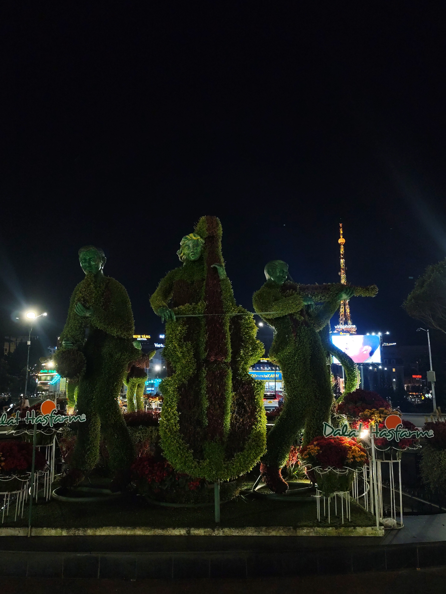 Three topiary statues depicting musicians stand on a concrete base at night. From left to right, the statues represent a musician holding a tambourine, a musician with a double bass, and a musician playing a violin. The statues are surrounded by flower beds with red and orange flowers. Signage reading "Dalat Hasfarm" is visible in front of the flower beds. In the background, streetlights illuminate the area, and a tall tower is visible. A large advertising screen displays a face. The location is Da Lat, Vietnam.