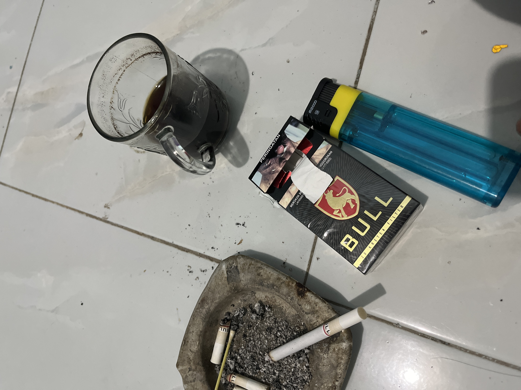 The image depicts a still life arrangement on a tiled floor, likely in a residential setting in Sumbernadi, Indonesia. In the foreground, a weathered ashtray contains several cigarette butts and ash, suggesting recent use. To the right of the ashtray, a pack of "BULL" brand kretek filter cigarettes is visible, partially opened to reveal a white tissue. Adjacent to the cigarette pack lies a blue and yellow disposable lighter. A clear glass mug filled with dark liquid, presumably coffee, sits further back and to the right, with some residue visible on the rim. The tiled floor appears to be light-colored with some scattered debris, including ash and dust. The overall scene conveys a sense of a casual, possibly solitary moment of relaxation or a break.