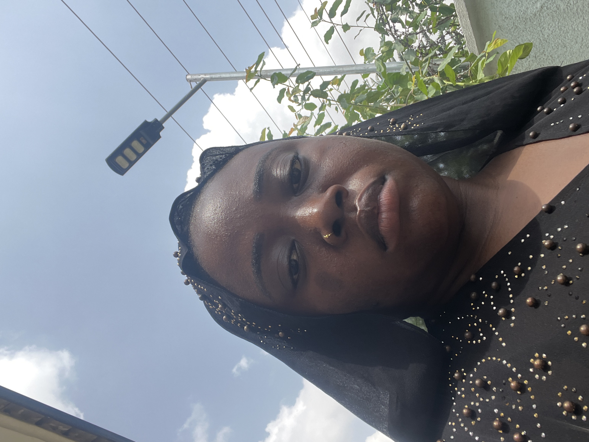 A close-up, low-angle portrait features a dark-skinned woman wearing a black garment adorned with small, gold-colored studs. She has a nose ring and is looking directly at the camera with a slight frown. The background includes a partly cloudy sky, utility wires, a black LED street light, and some green foliage. The exact location in Abuja, Nigeria cannot be definitively determined from the information present.