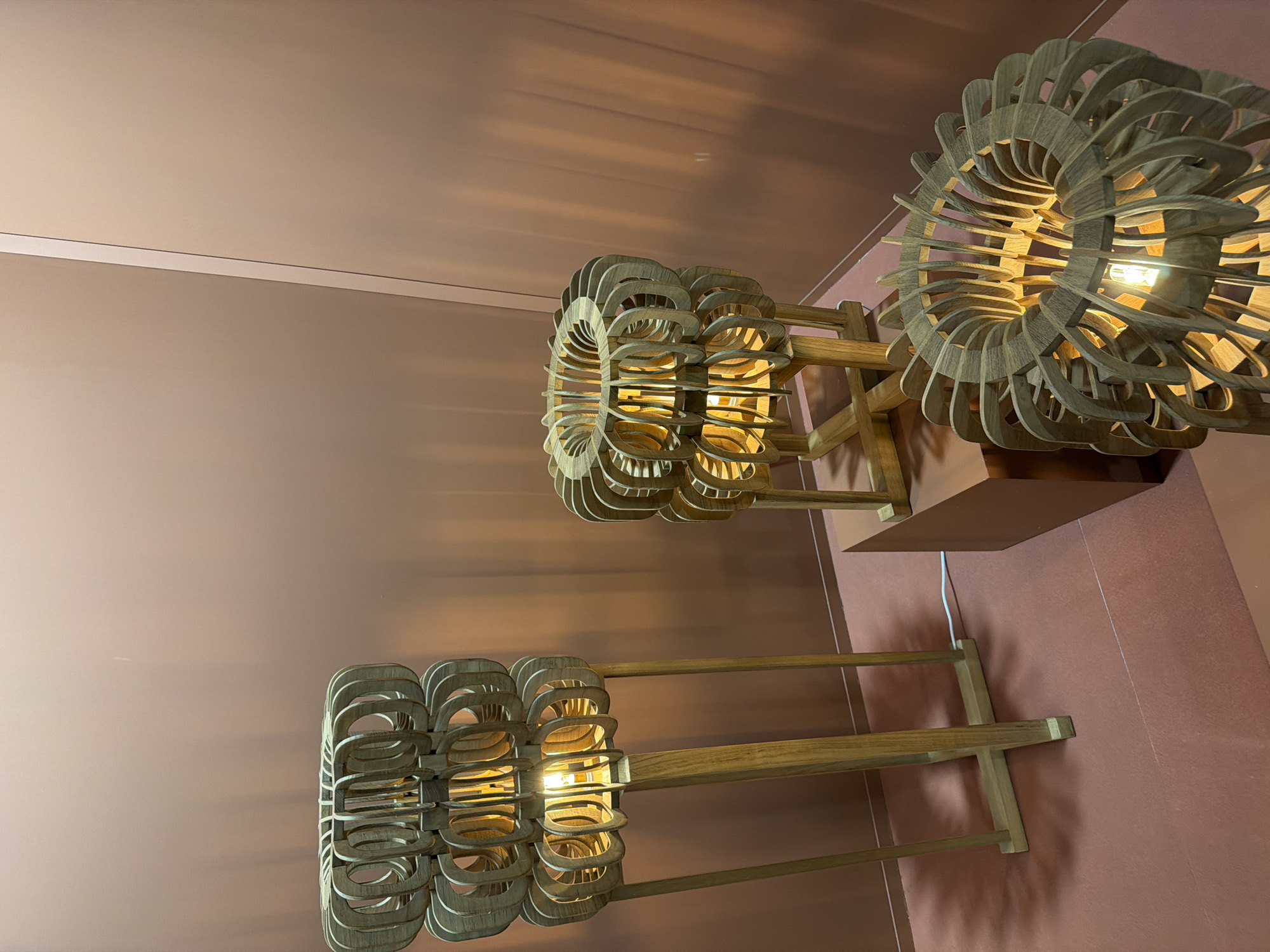 The image showcases an indoor setting featuring three decorative wooden light fixtures against a warm, muted background. The walls and floor appear to be a consistent terracotta or dusky rose color, creating a minimalist and cohesive environment.
Each of the three main objects is an illuminated structure crafted from numerous curved, light-toned wooden slats arranged in layered, concentric patterns, resembling abstract floral or geometric designs. A warm-glowing light bulb is positioned within the center of each fixture, casting intricate shadows and warm light onto the surrounding surfaces. These fixtures are supported by simple wooden stands or frames, elevating them.
In the foreground on the left, the lower section of one such illuminated piece is visible. Another complete fixture stands prominently in the mid-ground, slightly to the left of the center. A third, similarly designed piece is positioned further back and to the right, appearing to rest on a low, color-matched pedestal. A thin white electrical cord is visible near the base of the rightmost fixture.
The ambient lighting is dim, emphasizing the warm glow of the lamps, which serve as the primary light sources. There is no activity taking place, indicating a static display. No people or visible text are present in the image. The scene suggests a focus on modern design, ambient lighting, or artistic exposition within a contemporary interior space.