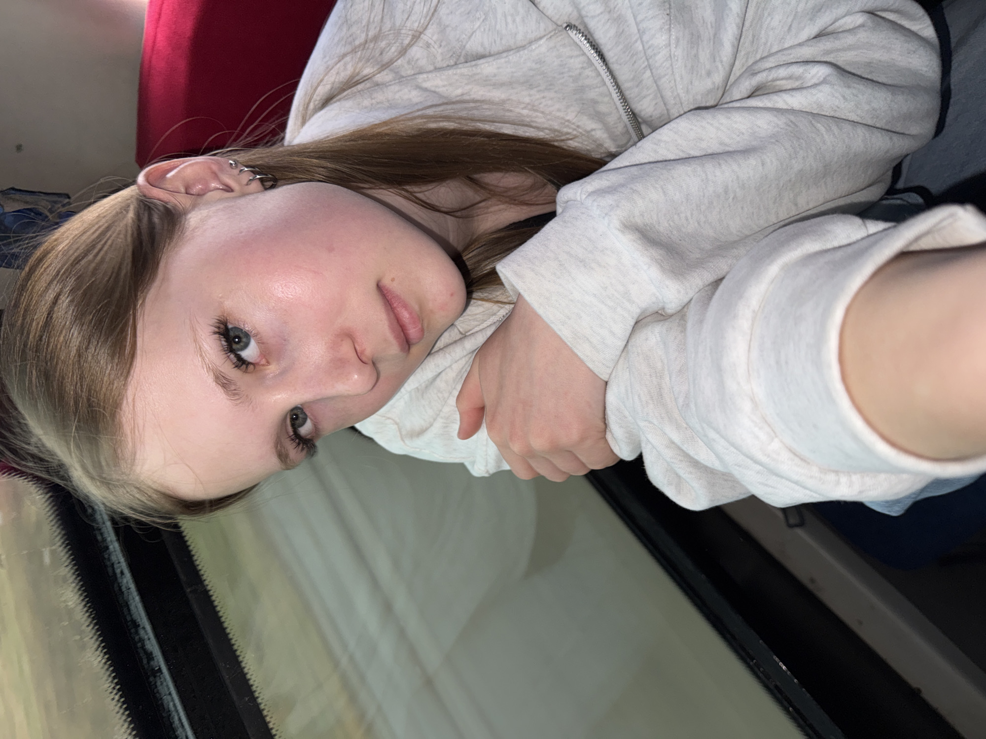 A young woman with light brown hair and fair skin is looking at the camera. She is wearing a light gray zip-up hoodie and appears to be resting her arm on a window. Part of a red seat is visible behind her head. The image does not contain enough information to determine the location in Froly, Russia.