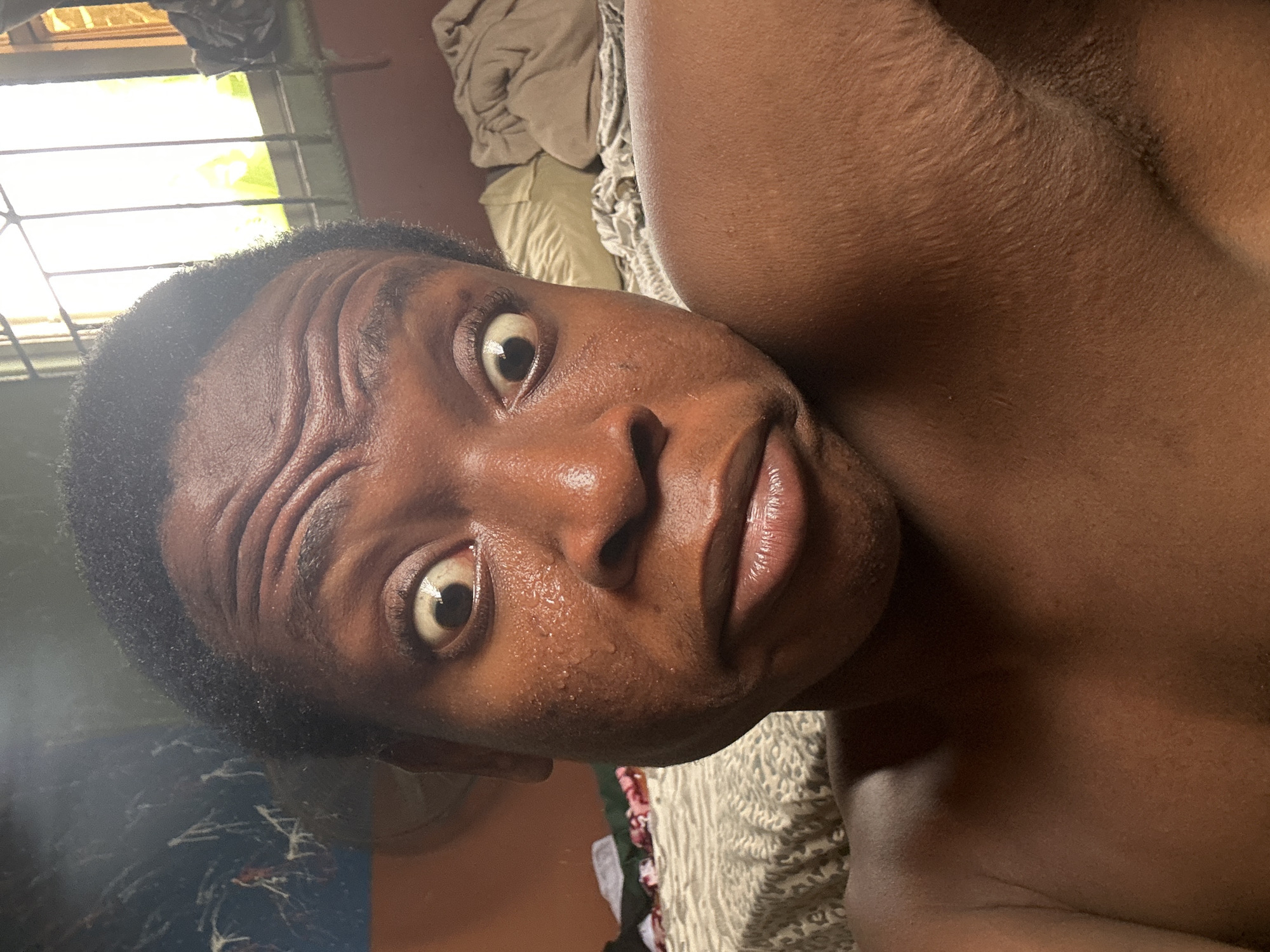 The image depicts a person lying down, facing upwards towards the camera. The person has dark skin, short dark hair, and is visibly sweating. Their eyes are wide open, and their forehead is wrinkled. The person is partially undressed, revealing a portion of their chest and upper body. The background includes a window with bars, a wall painted in a dull color, and bedding. The location is identified as Unknown, Unknown.