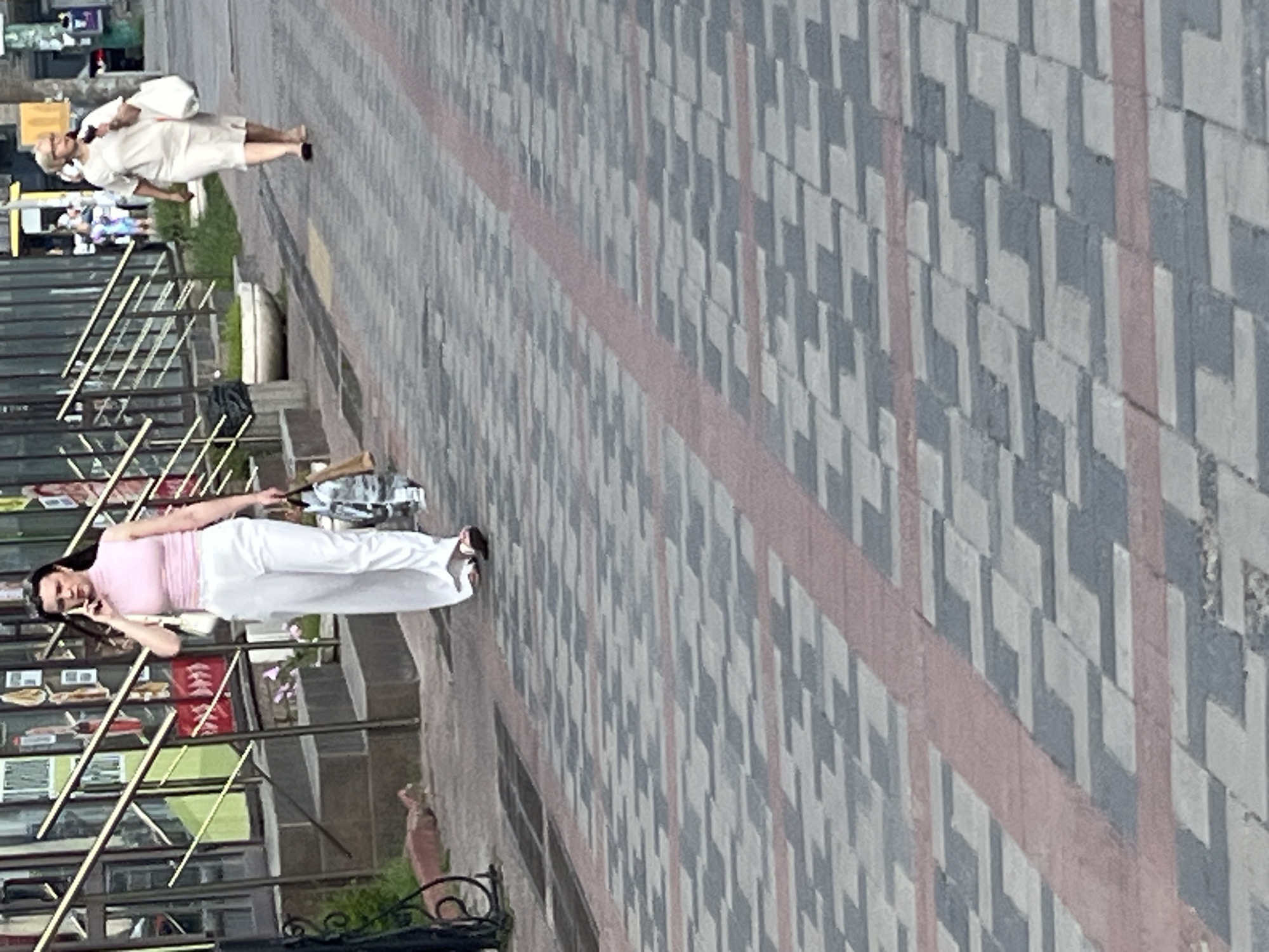 A woman, wearing a pink top and white pants, is talking on a mobile phone while walking along a sidewalk. She appears to be alone, and her posture suggests she is engaged in a conversation. In the background, another woman in a white dress and blonde hair is also walking. The scene is set on a paved sidewalk with a geometric pattern of grey and white tiles, bordered by a pink stripe. There are railings and structures, possibly shop fronts, visible to the left of the sidewalk. The lighting suggests it is daytime, and the weather appears to be clear. The overall setting is an urban environment, likely a street or pedestrian area in Dnipro, Ukraine. No text is clearly legible in the image.
