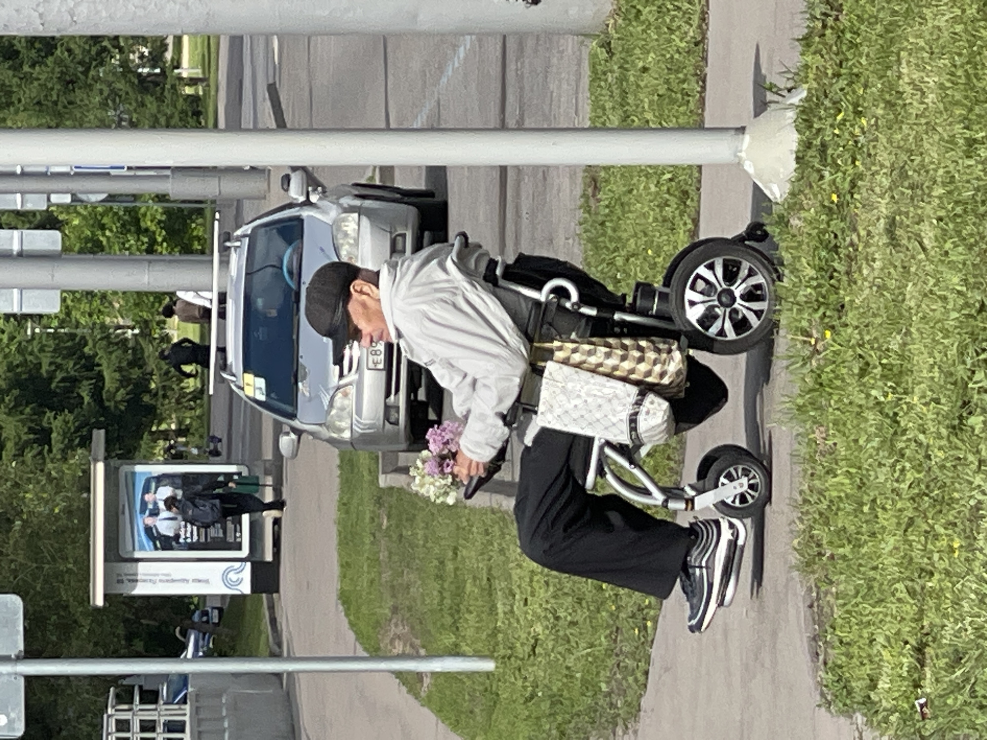 A senior man is riding an electric scooter on a sidewalk next to a street. He is wearing a light gray jacket, dark pants, and black and white striped sneakers. The man is holding a bouquet of lilac flowers in his left hand. The scooter has a basket attached to the front. A light silver car is parked on the street next to the sidewalk. Behind the car, there is a bus stop with an advertisement. In the background, there are trees and buildings. The photo was taken in Moscow, Russia based on the license plate on the parked car.
