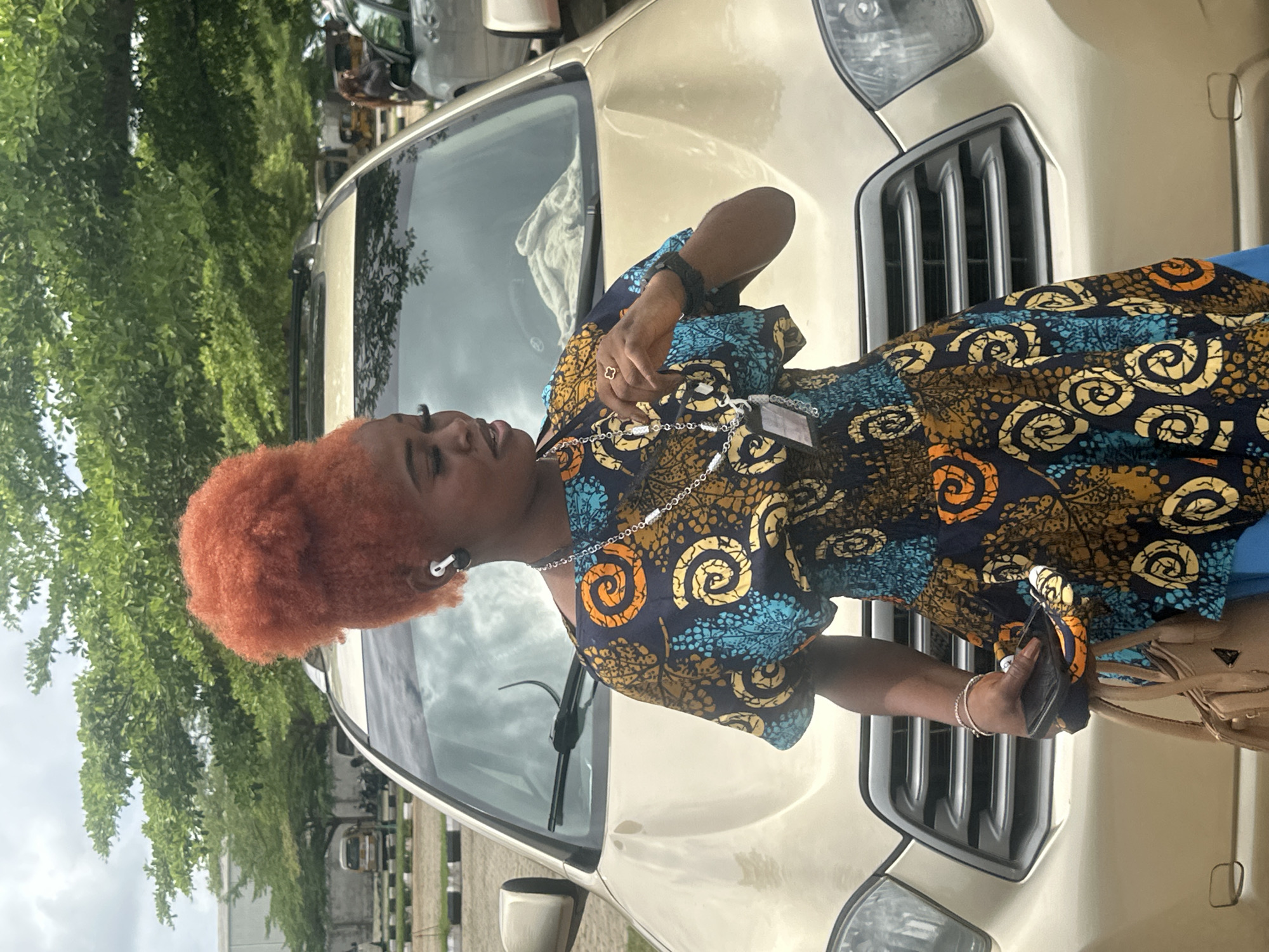 A young Black woman with bright orange, voluminous hair stands in front of a gold SUV. She is wearing a patterned dress with blue, orange, and yellow swirls. She has a silver necklace with multiple pendants and an earbud in her left ear. In her right hand, she holds a black item and a light brown purse is slung over her left arm. The background shows trees and a cloudy sky, suggesting an outdoor setting in Itoga, Nigeria.