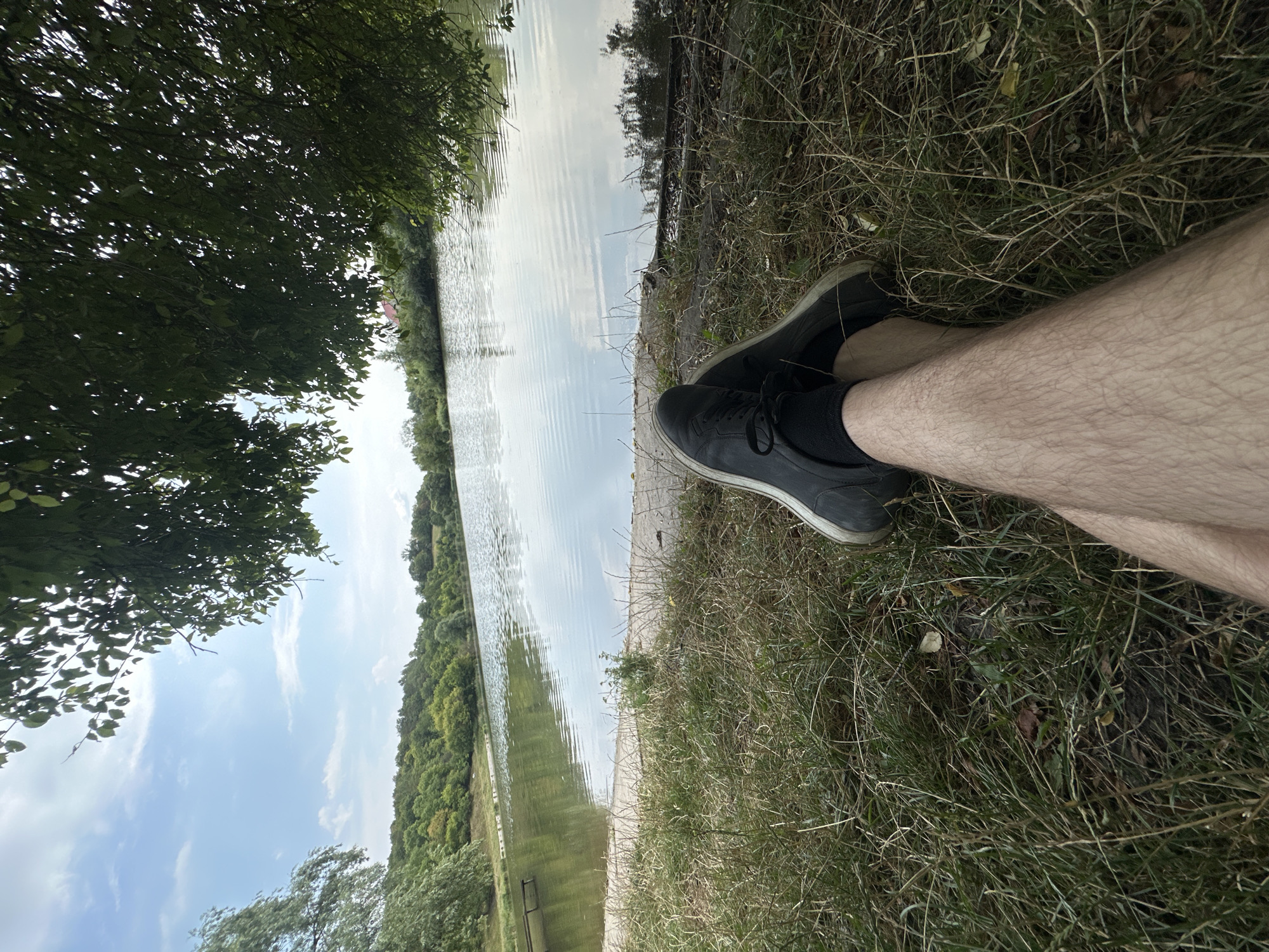 A first-person perspective shot shows a person's legs and feet crossed at the ankles, resting on a grassy embankment. The individual is wearing dark sneakers and black socks. The scene is set by a calm lake, reflecting the sky and the lush green trees that line its opposite shore. The foliage of a large tree with dark green leaves hangs over the right side of the frame. The location is the lake in Makiv, Ukraine.